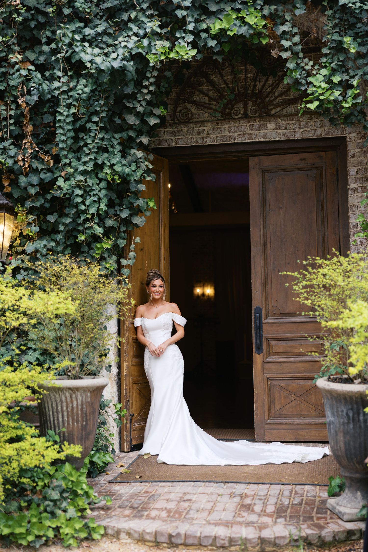 Radiant bride in off-shoulder fitted white gown stands in ivy-covered stone doorway before her wedding ceremony.