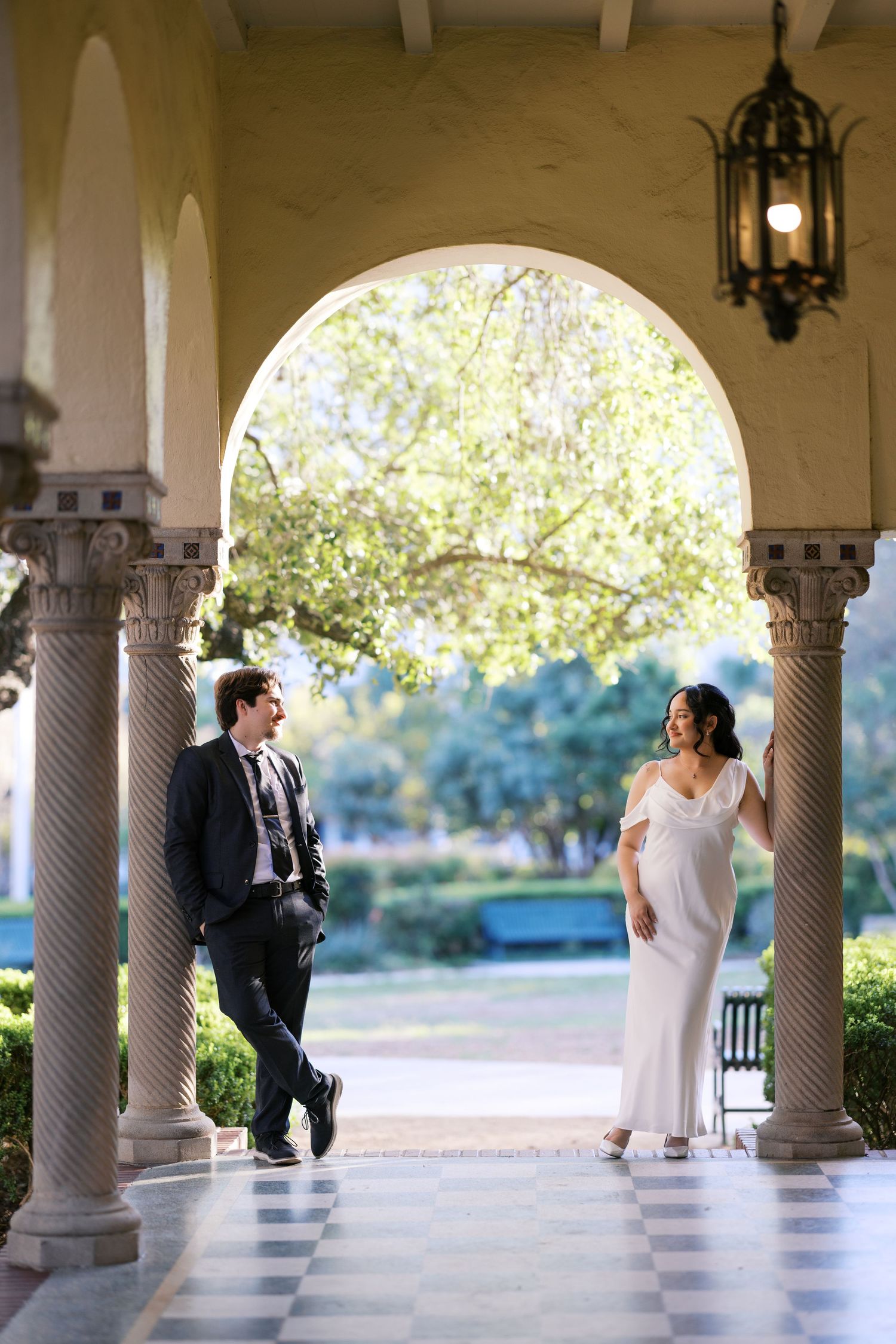 A bride and groom share a romantic moment under elegant arched columns at a classic venue with lush gardens.