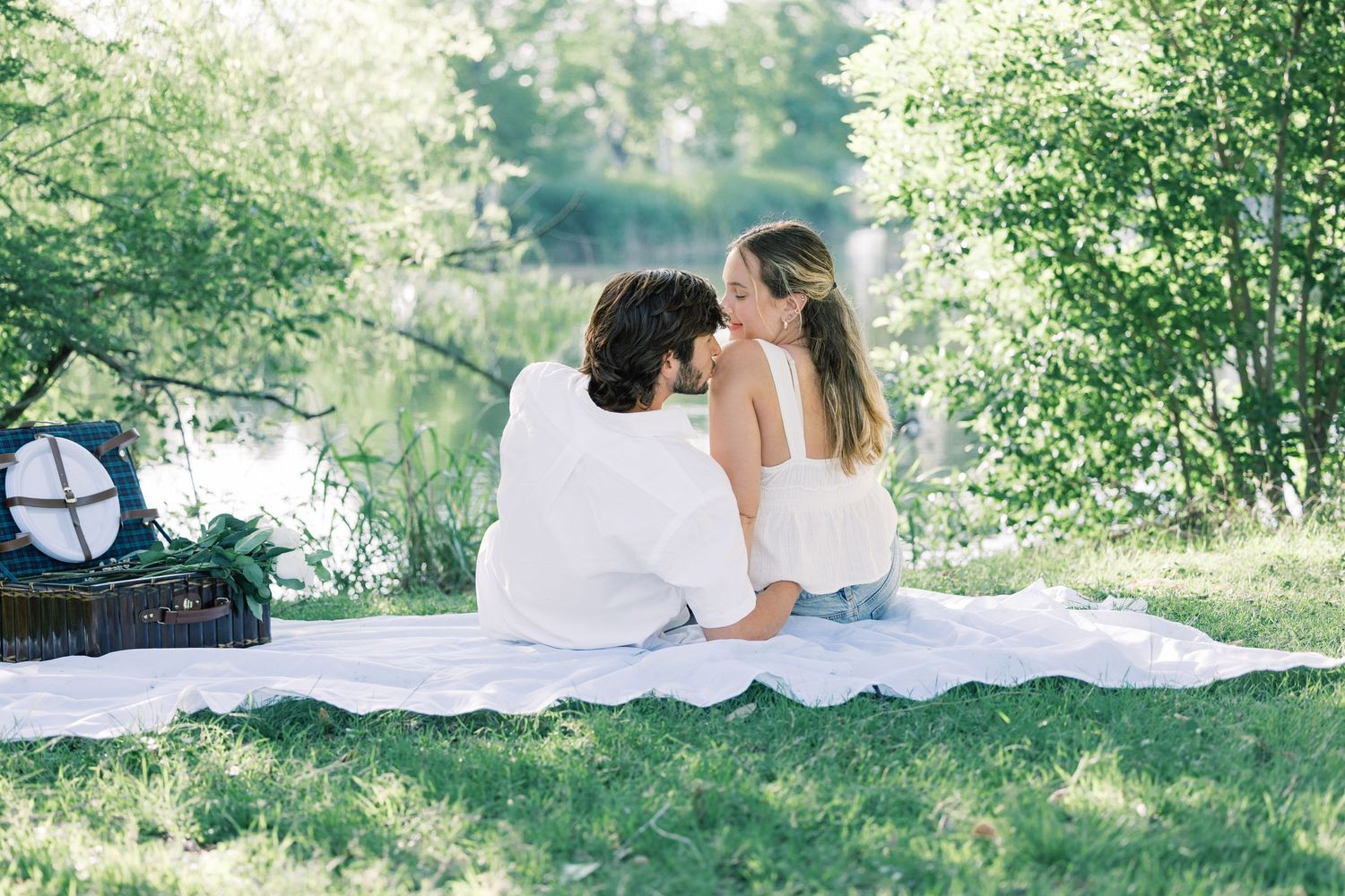 A romantic picnic scene unfolds by a lake with trees providing natural shade.