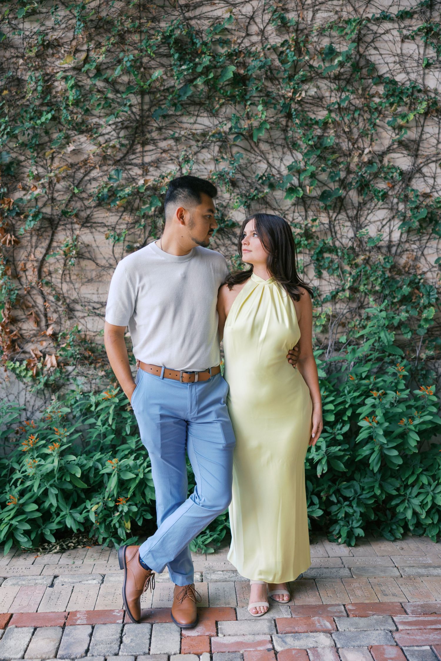 A couple poses together against a vine-covered wall wearing casual summer outfits.