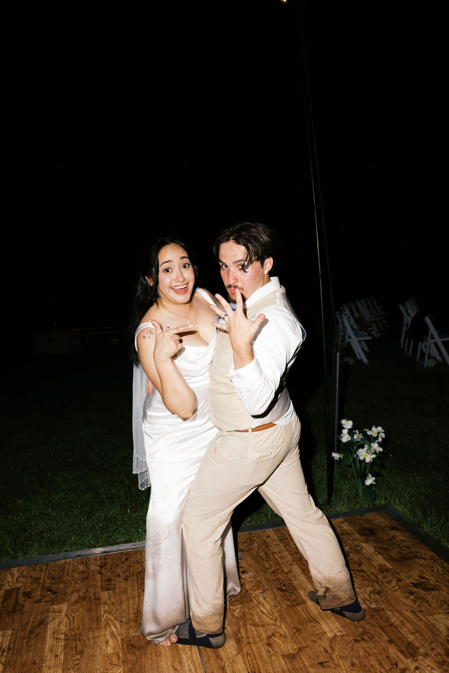 Newlywed couple poses playfully on dance floor at night, showing off wedding rings at outdoor reception.