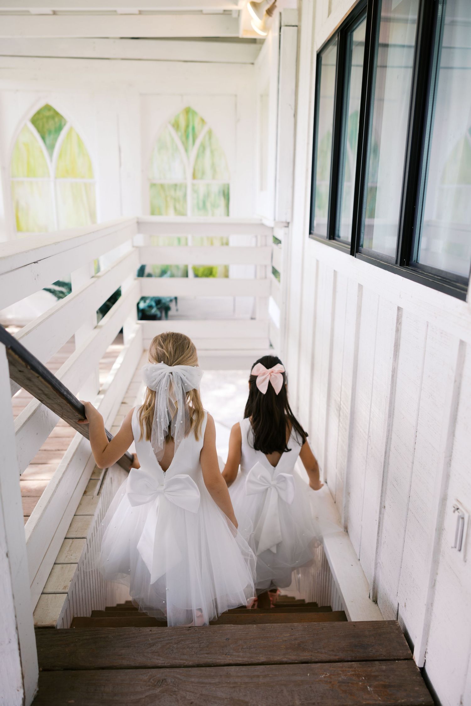 Two flower girls in white tulle dresses with bows climb wooden stairs inside a white chapel.