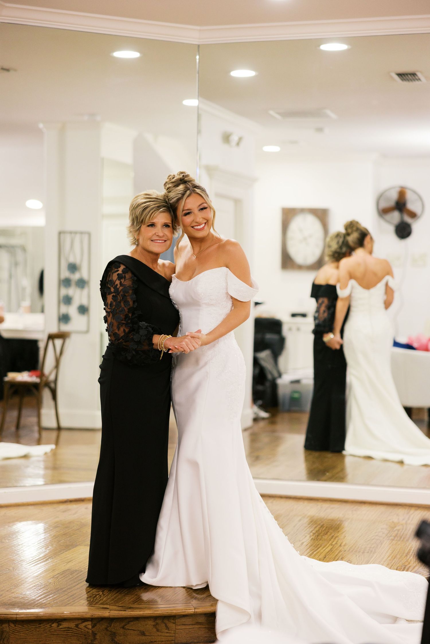 Bride in off-shoulder white wedding gown posing with mother in black dress in bridal boutique.