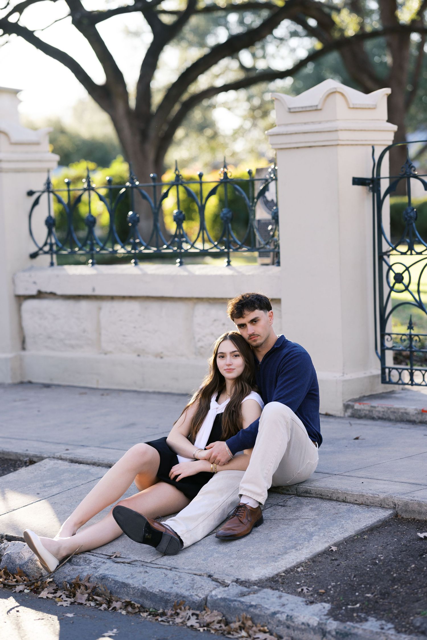 Young couple sitting together on steps near ornate iron gate, posing for an outdoor portrait photo.