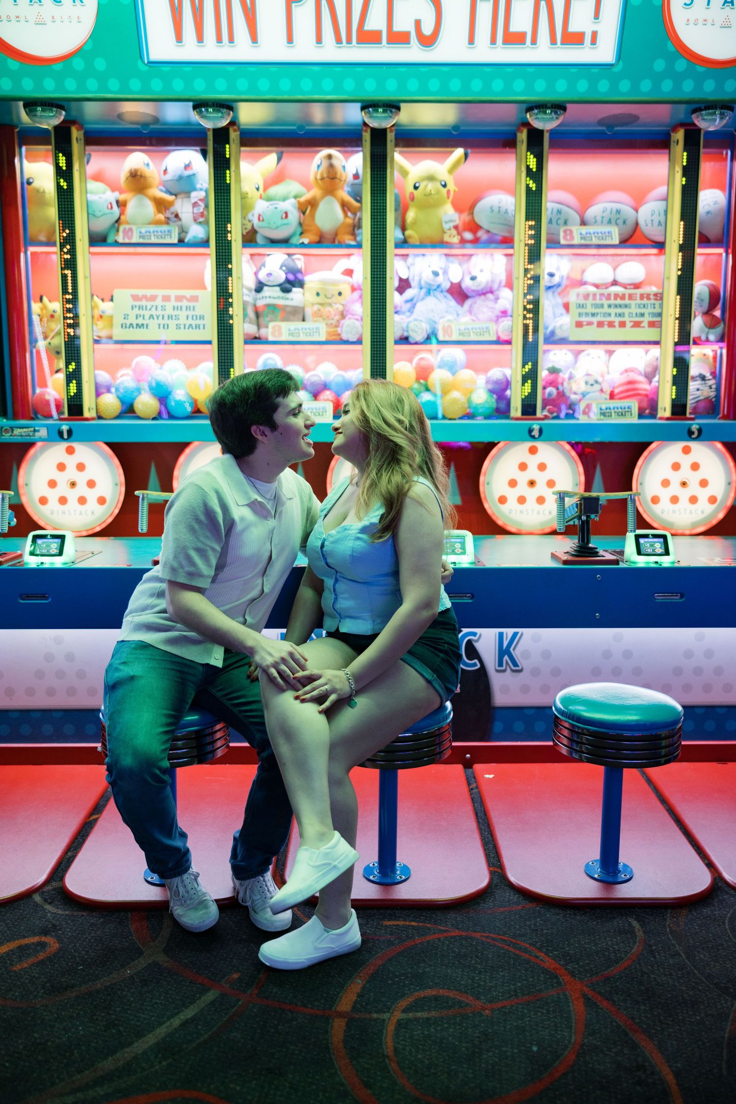 Couple sitting at an arcade game counter with colorful prize displays and neon lights in the background.