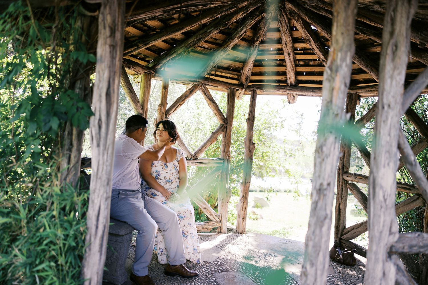 A couple shares an intimate moment in a rustic wooden gazebo surrounded by lush greenery.