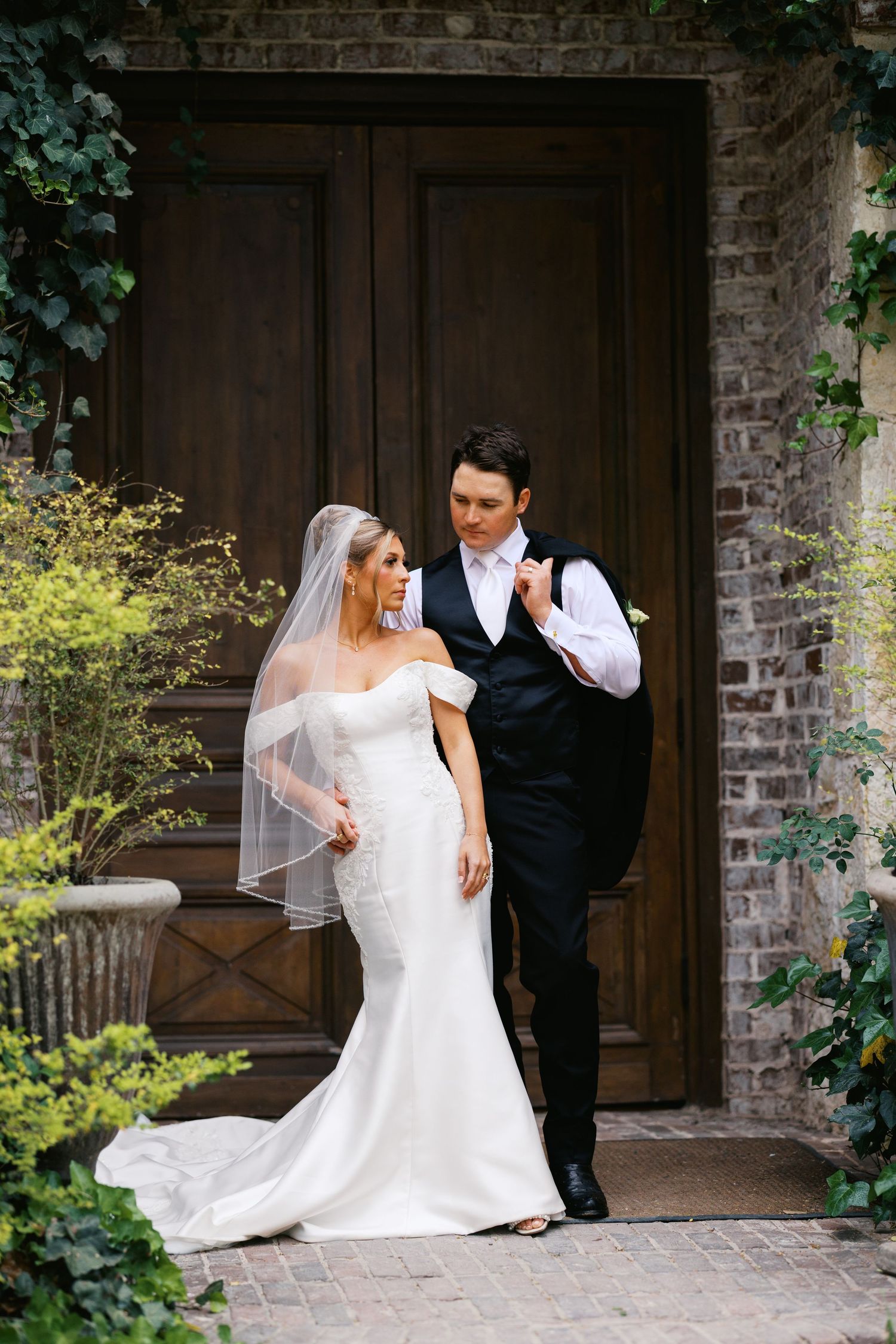 Elegant bride and groom pose together in front of ivy-covered brick wall and ornate wooden doors.