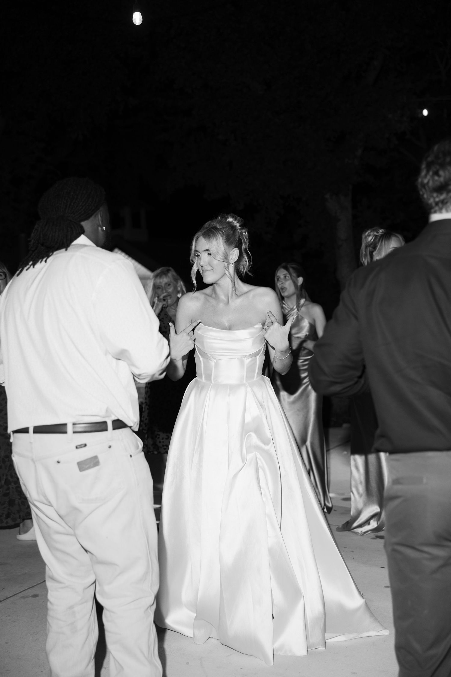 Black and white photo of a bride in a strapless ball gown dancing with guests at an outdoor wedding reception.