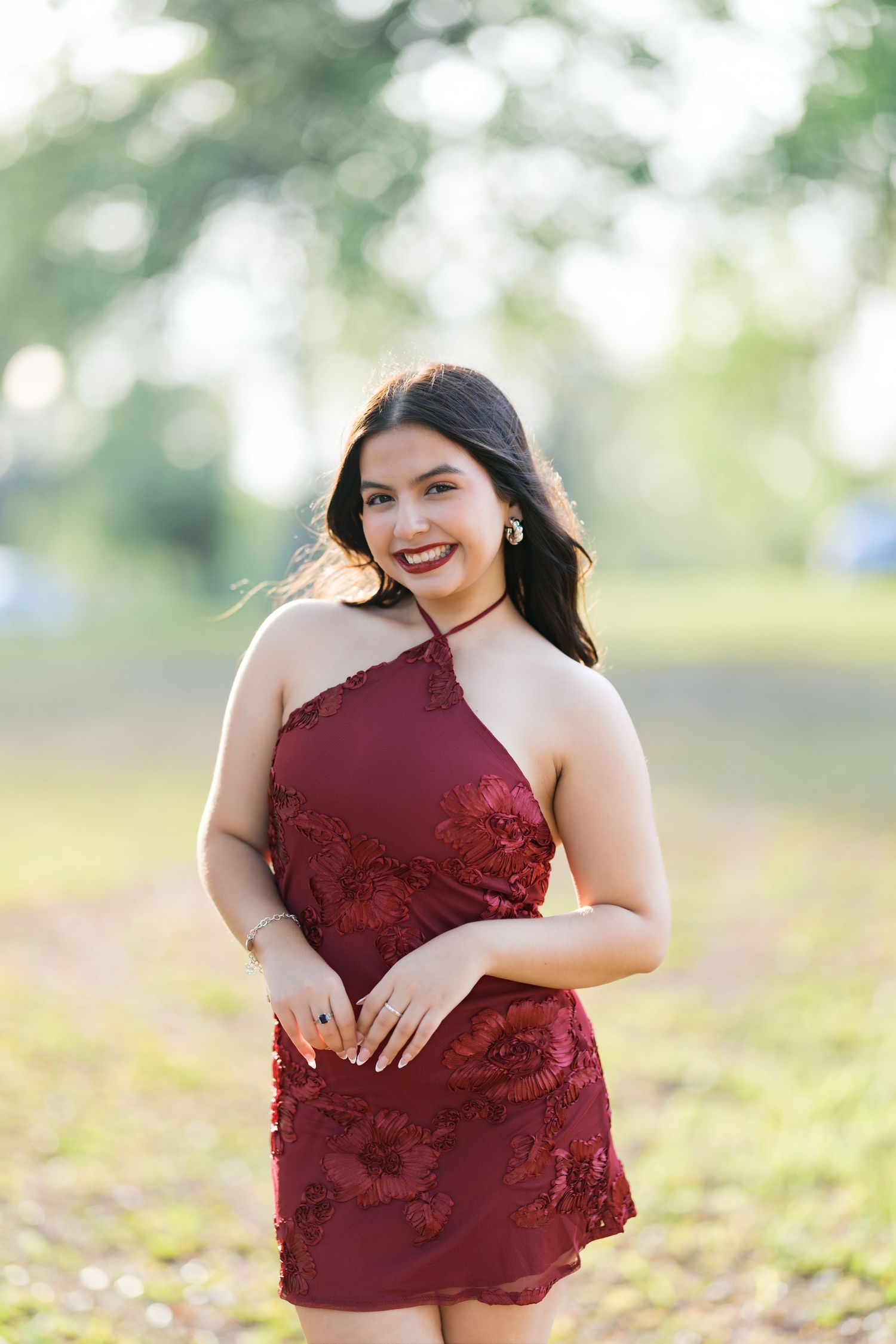 Young woman in a burgundy floral halter dress smiling outdoors in a sunlit park setting.