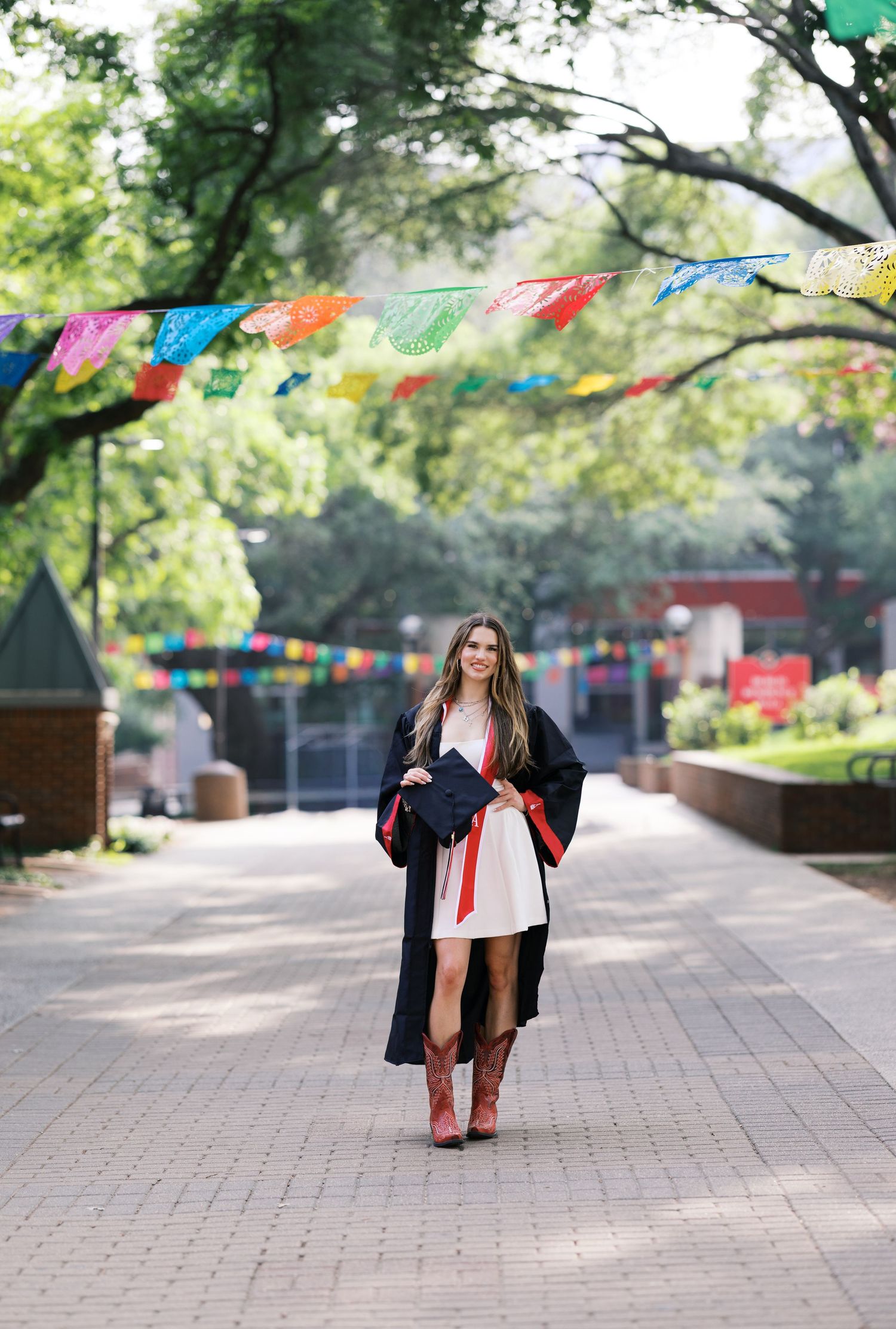 Female graduate in white dress and red boots wearing graduation gown walks on brick path with colorful flags.