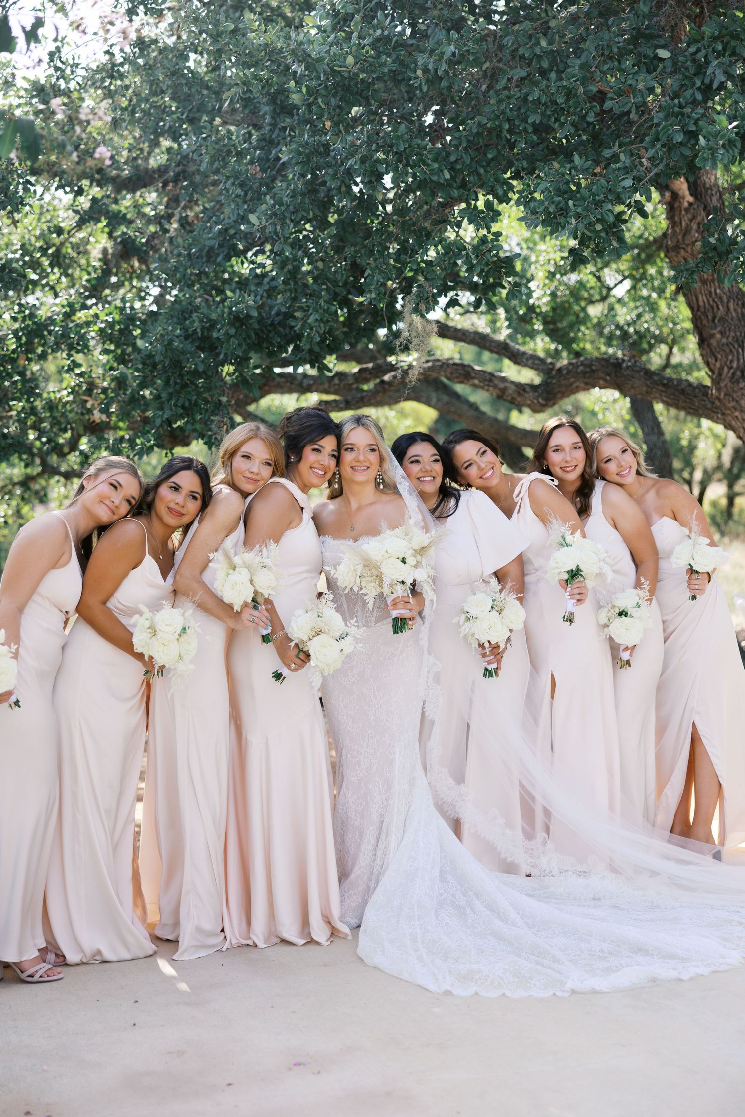 Wedding party in matching blush gowns gather for a joyful group photo outdoors.
