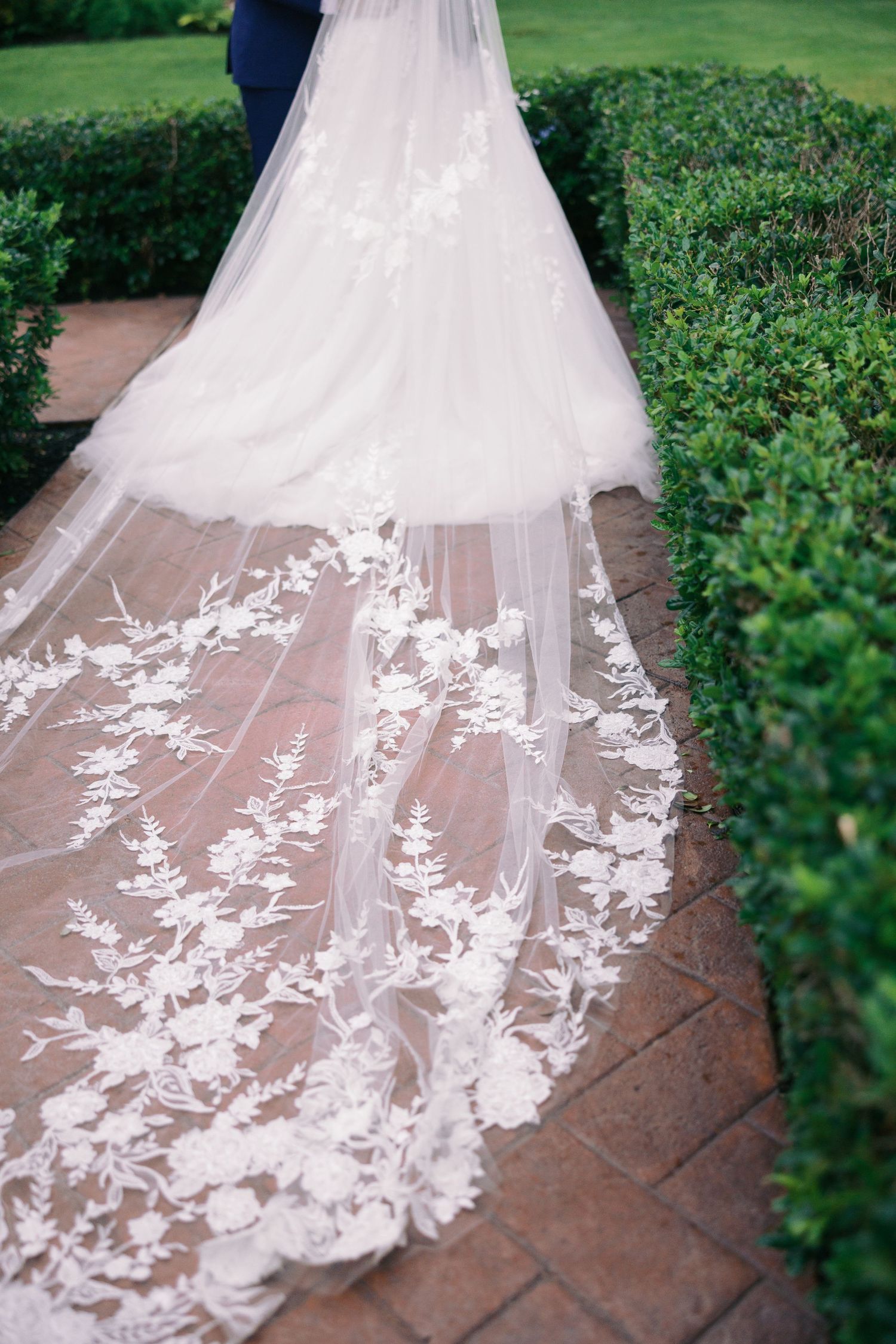 Close-up details of an ornate lace wedding dress train on terracotta tiles surrounded by greenery.