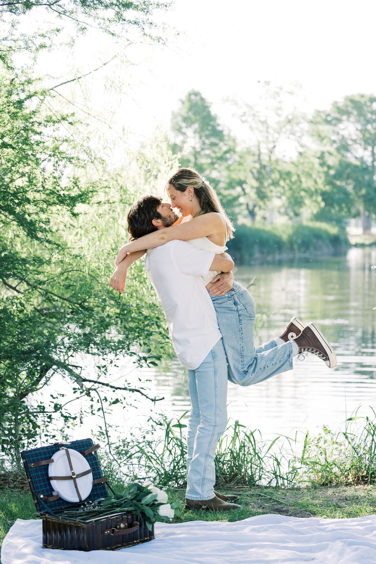 A romantic couple embracing by a lake with a picnic blanket and vintage record player in a dreamy outdoor setting.