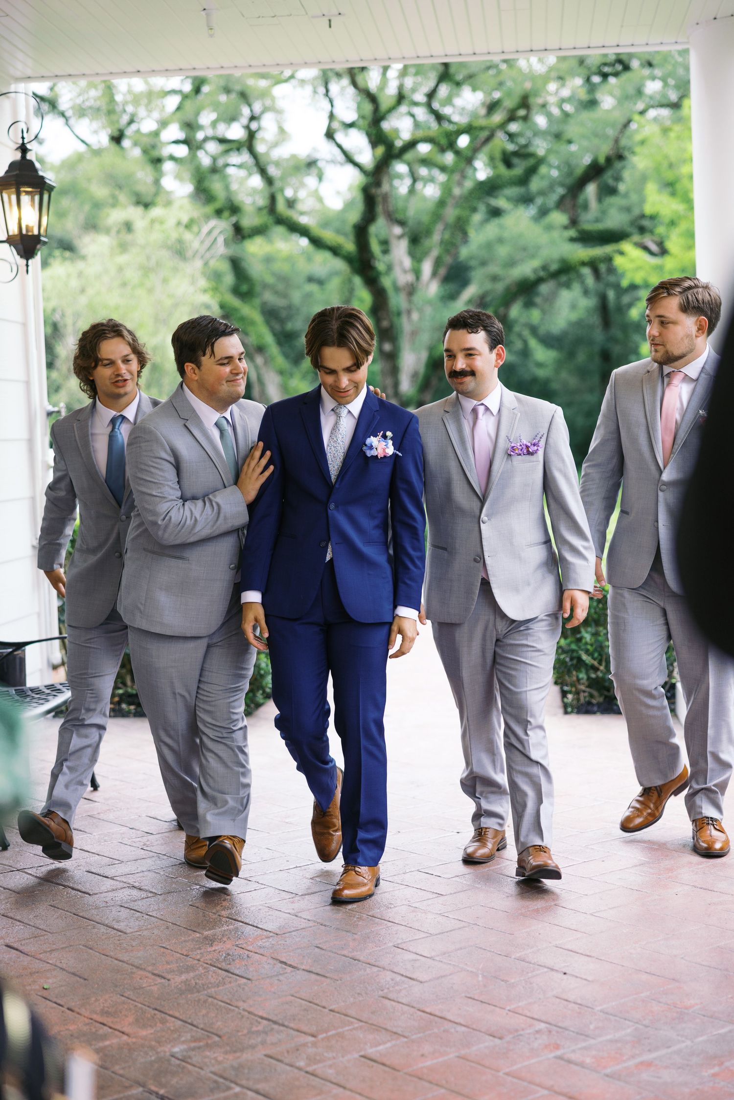 Groomsmen in coordinated gray suits and one in navy blue walking together on a brick porch with greenery in background.