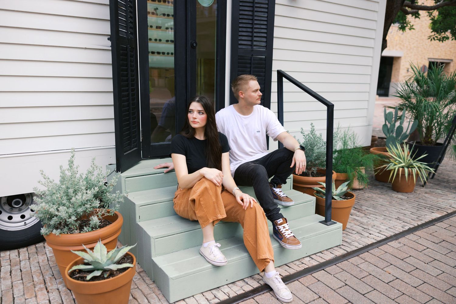 Casual couple sitting together on front porch steps with potted plants.