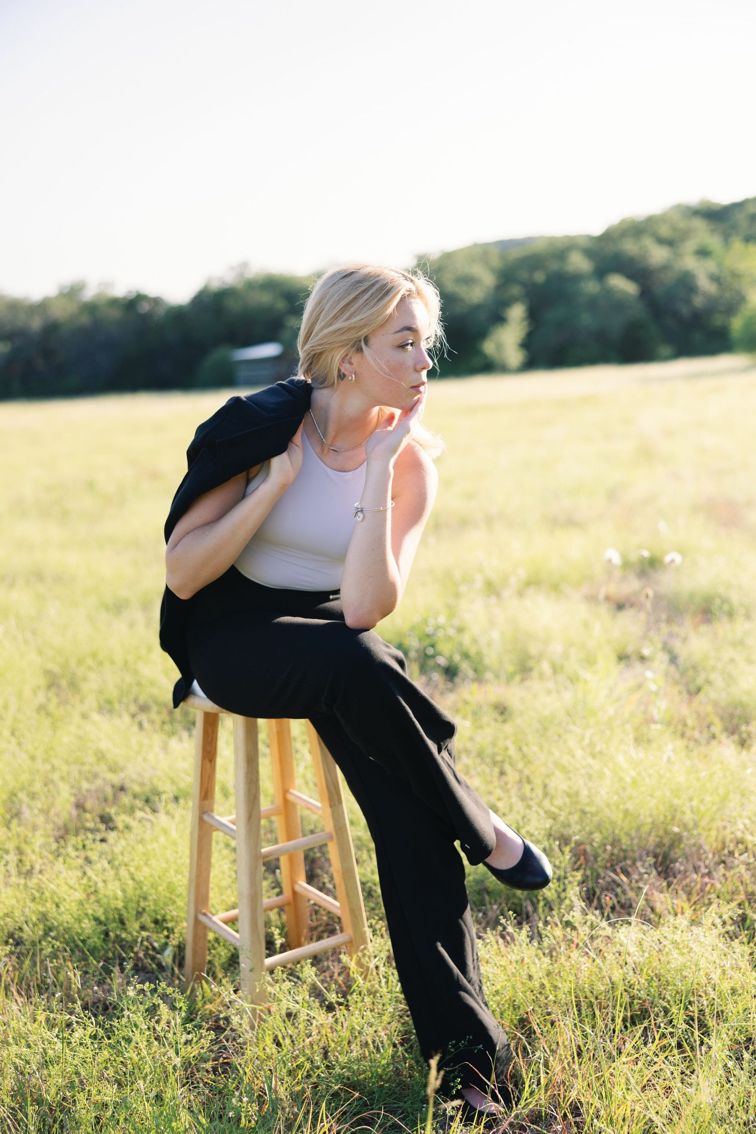 Dynamic poses on wooden stool in field wearing black blazer and white tank top.