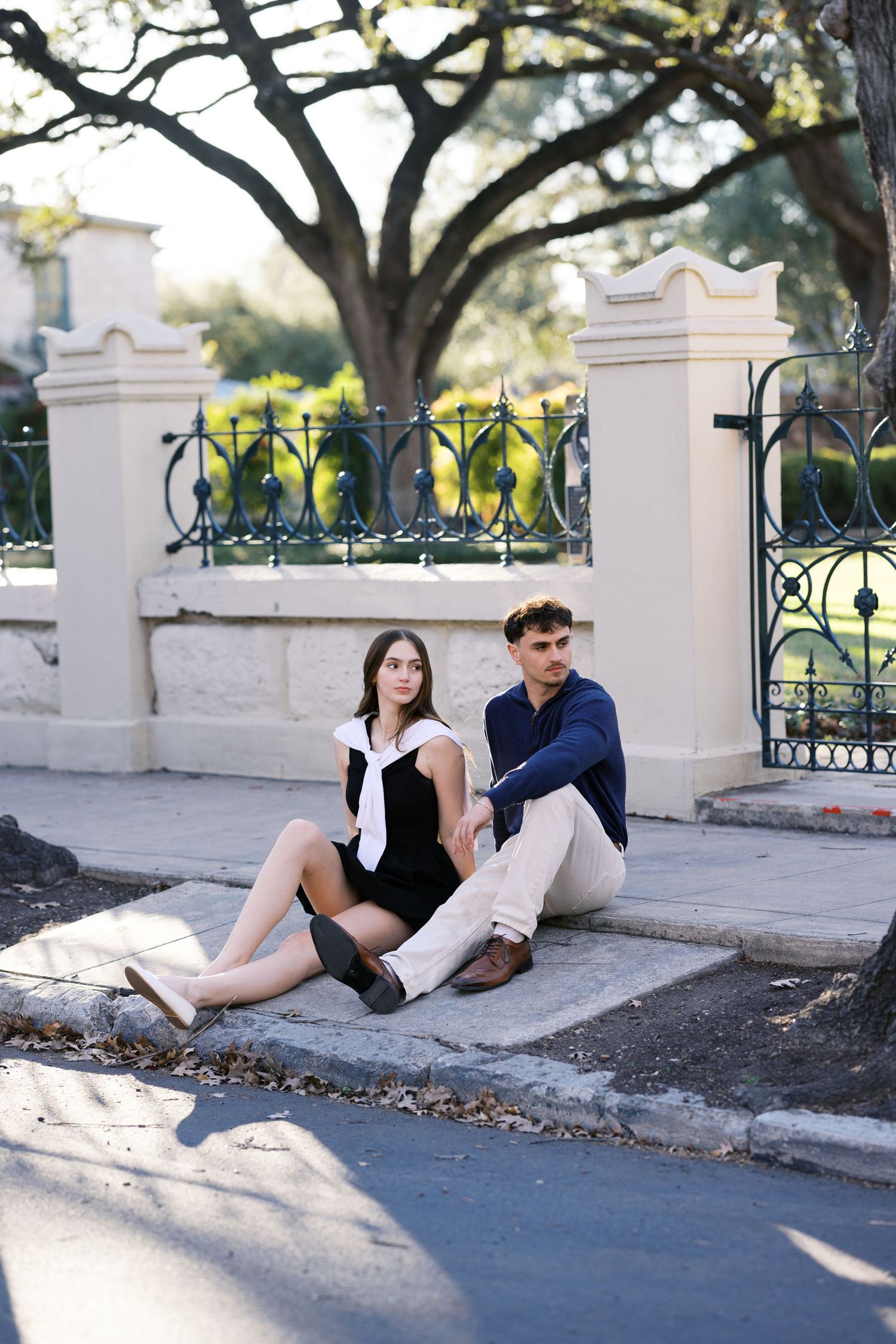 A young couple sits together on a curb near an ornate iron fence and stone pillars, surrounded by trees.