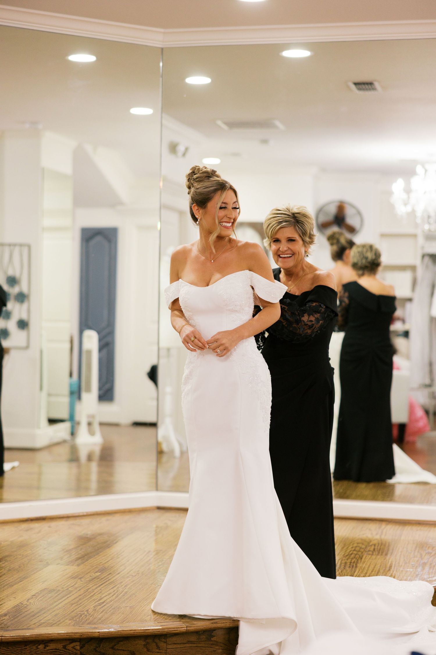 Bride in white off-shoulder mermaid gown smiles with woman in black dress in bridal boutique.
