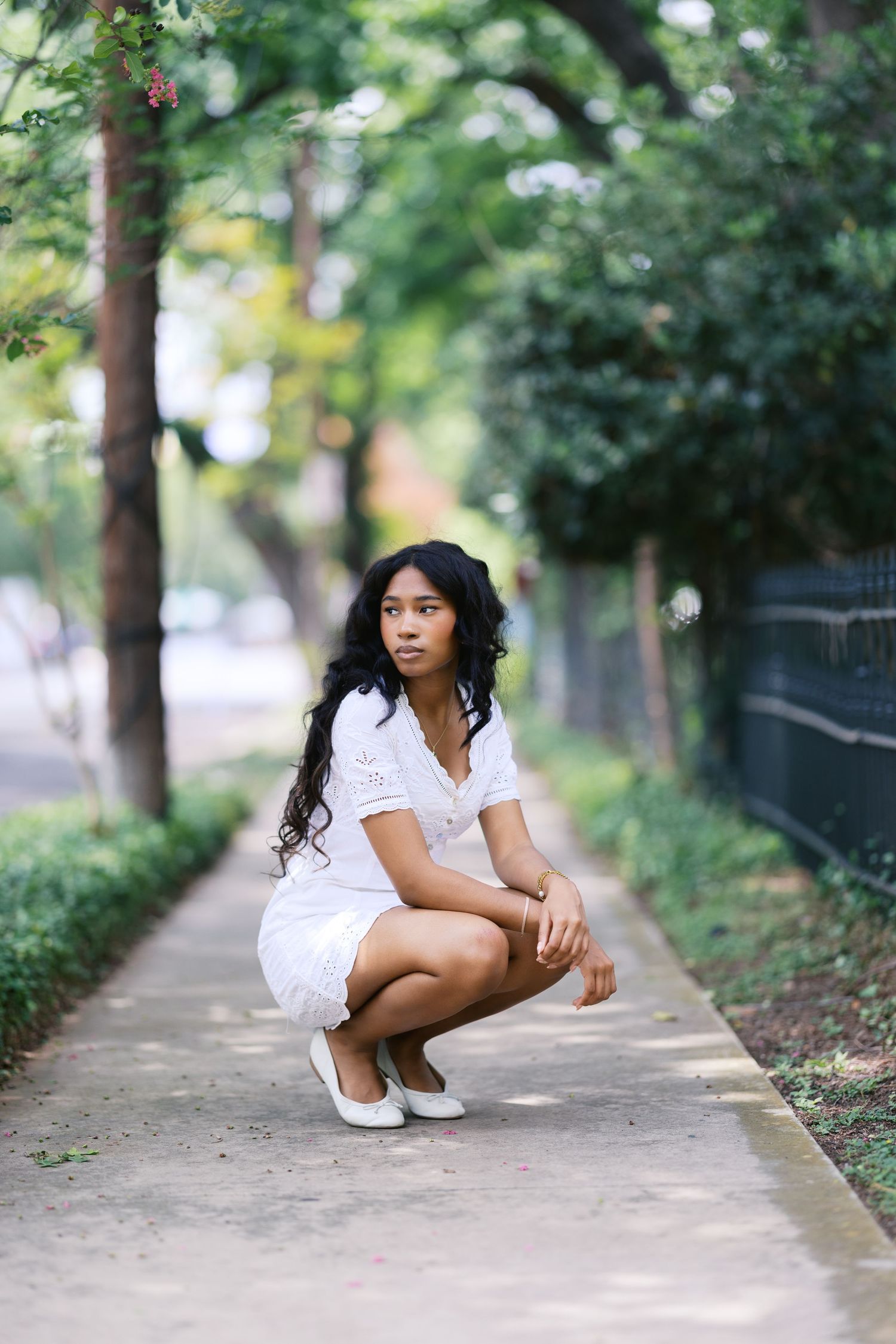 Young Black woman in white eyelet dress crouching on urban sidewalk surrounded by trees.