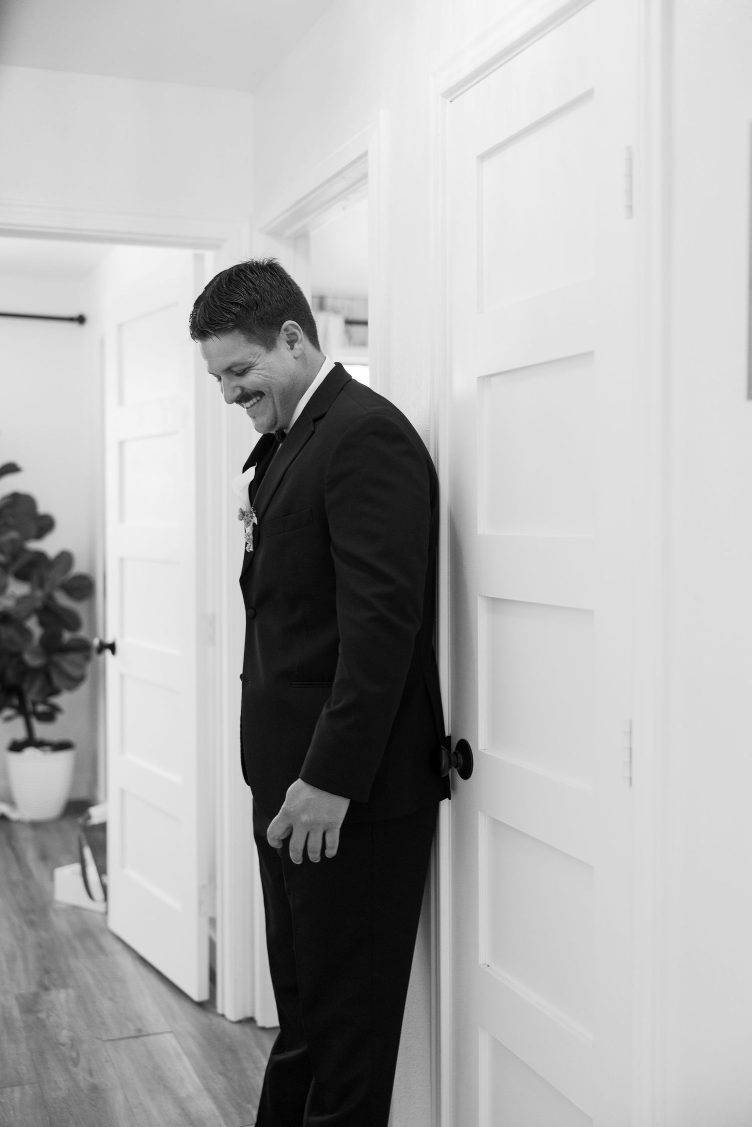 Groom in black tuxedo smiling while leaning against white door before wedding ceremony.