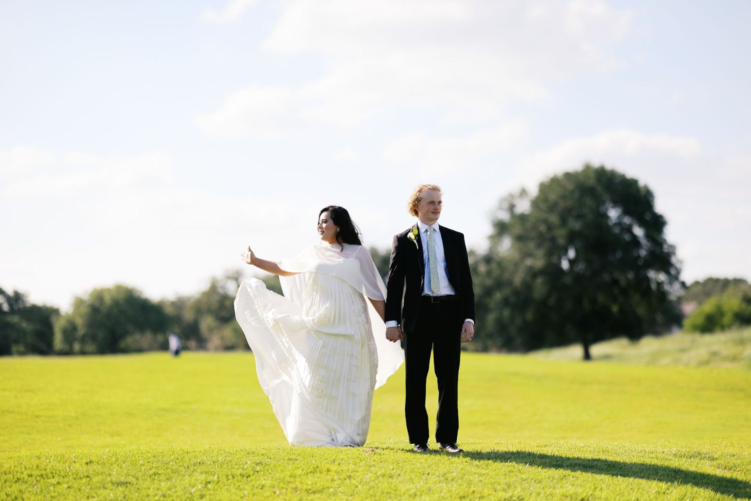 Bride in flowing white gown and groom in black tuxedo hold hands on lush green lawn under blue sky.