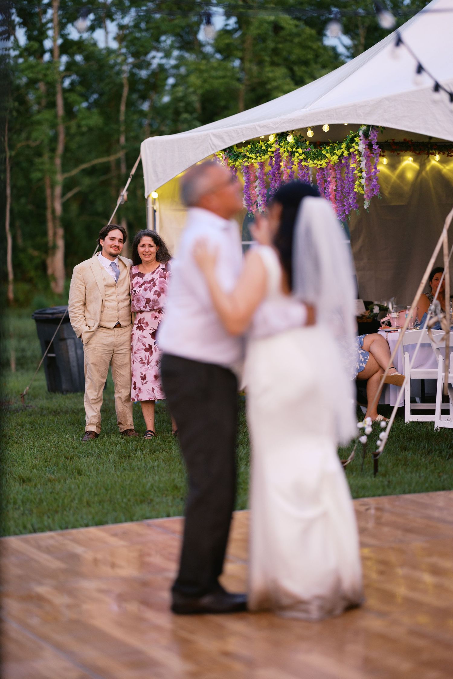 Bride and older man share first dance outdoors under lit tent while guests watch at wedding reception.