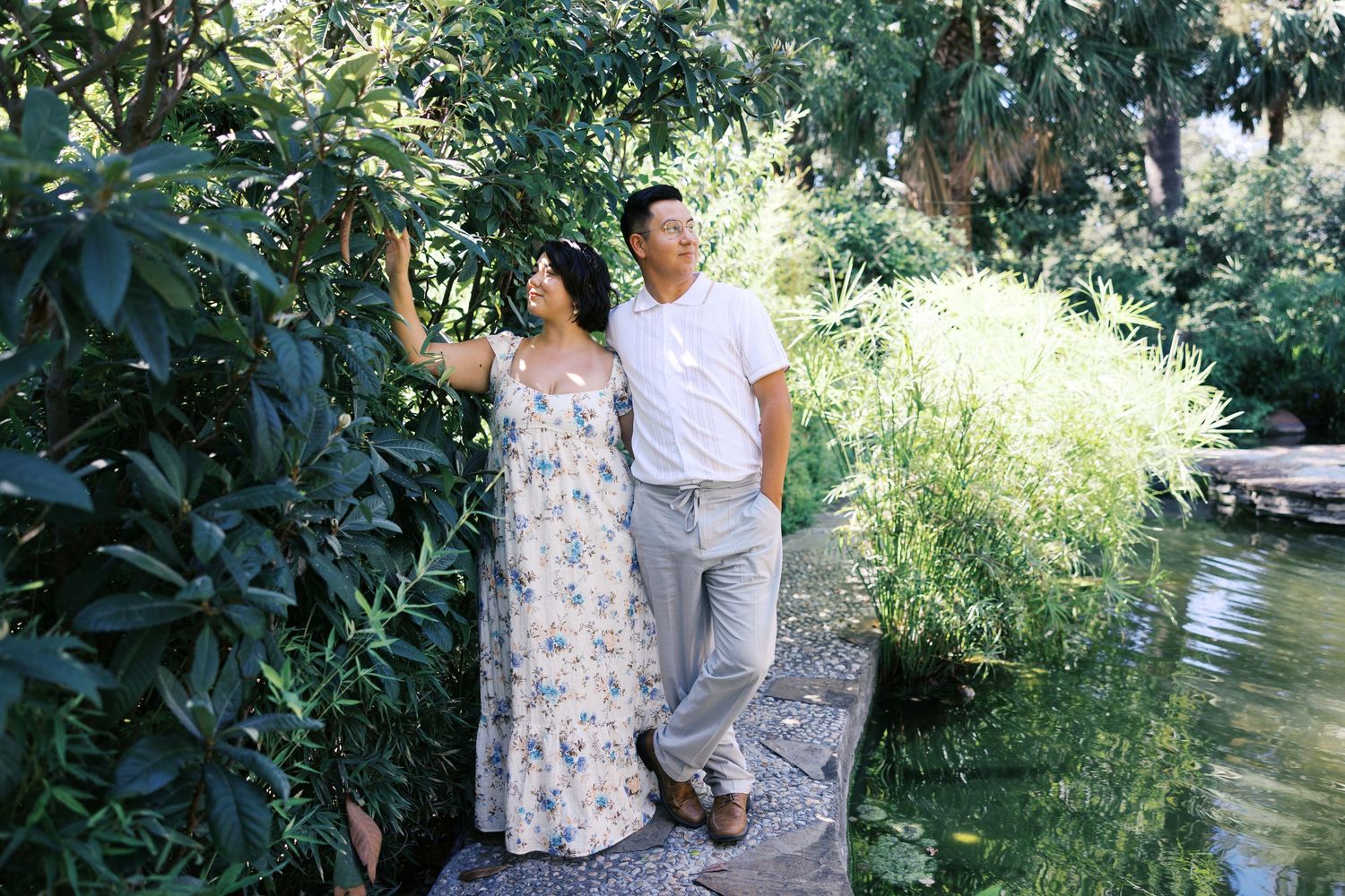 A playful garden photoshoot with a couple posing against lush foliage near a pond.