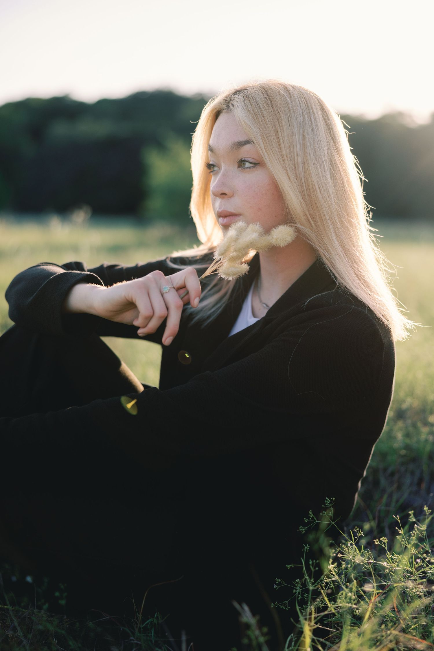 Portrait of a blonde woman in a black jacket posing thoughtfully in a sunlit field at golden hour.