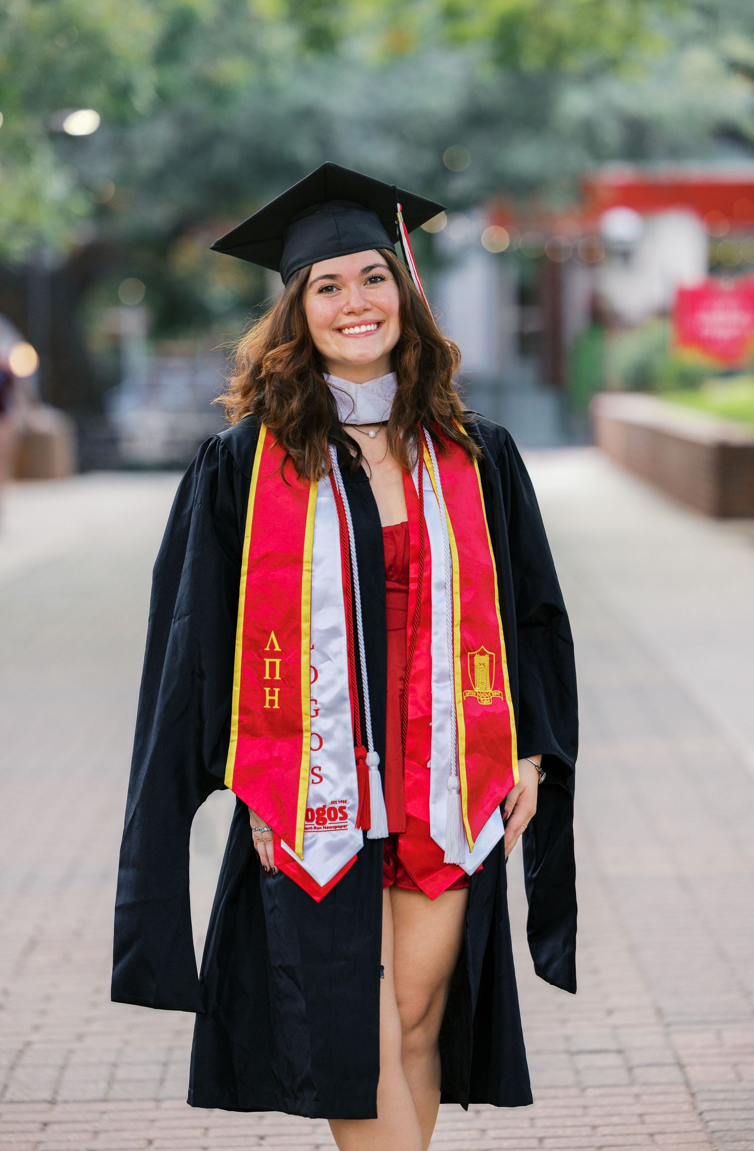 Graduate wearing academic regalia with red stoles standing on campus walkway.