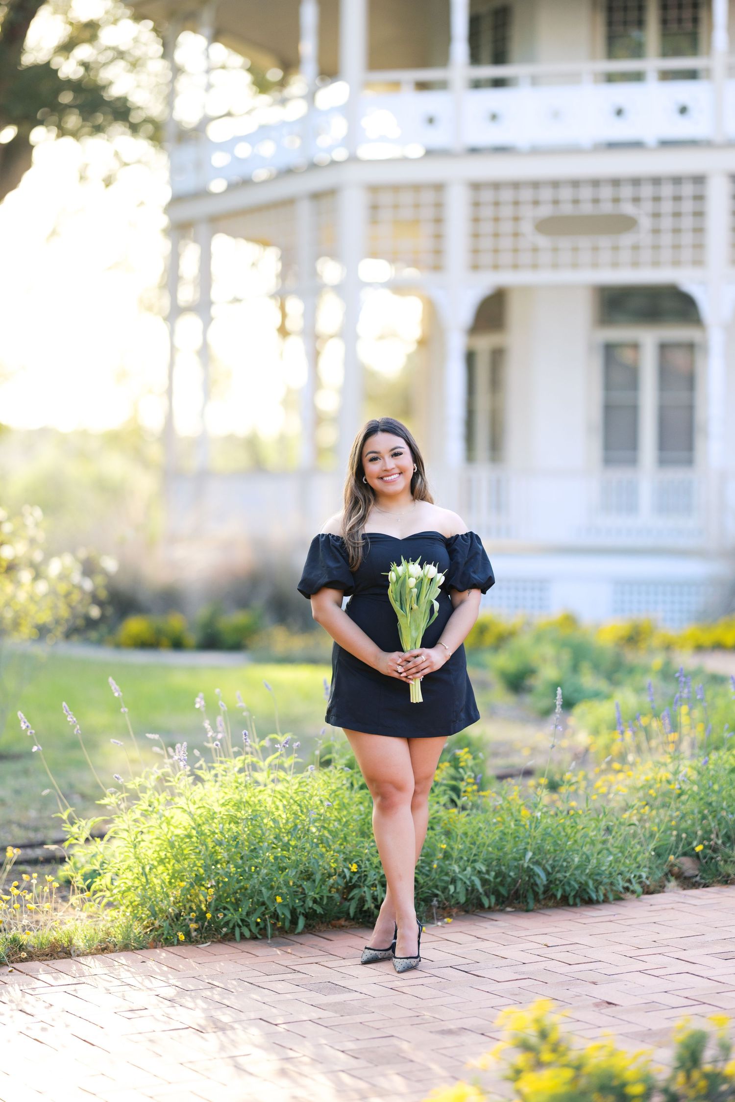 Young woman in black off-shoulder mini dress holding white flowers, smiling on brick path near white Victorian gazebo.