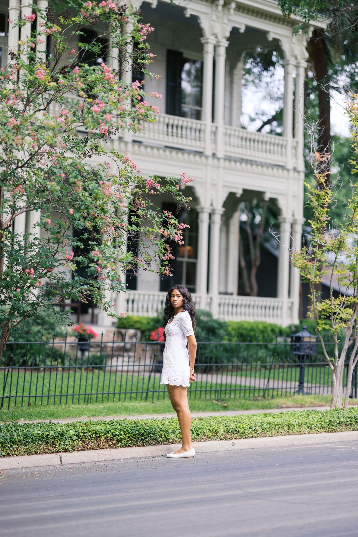 Young Black woman in white dress standing on street near historic Victorian mansion.