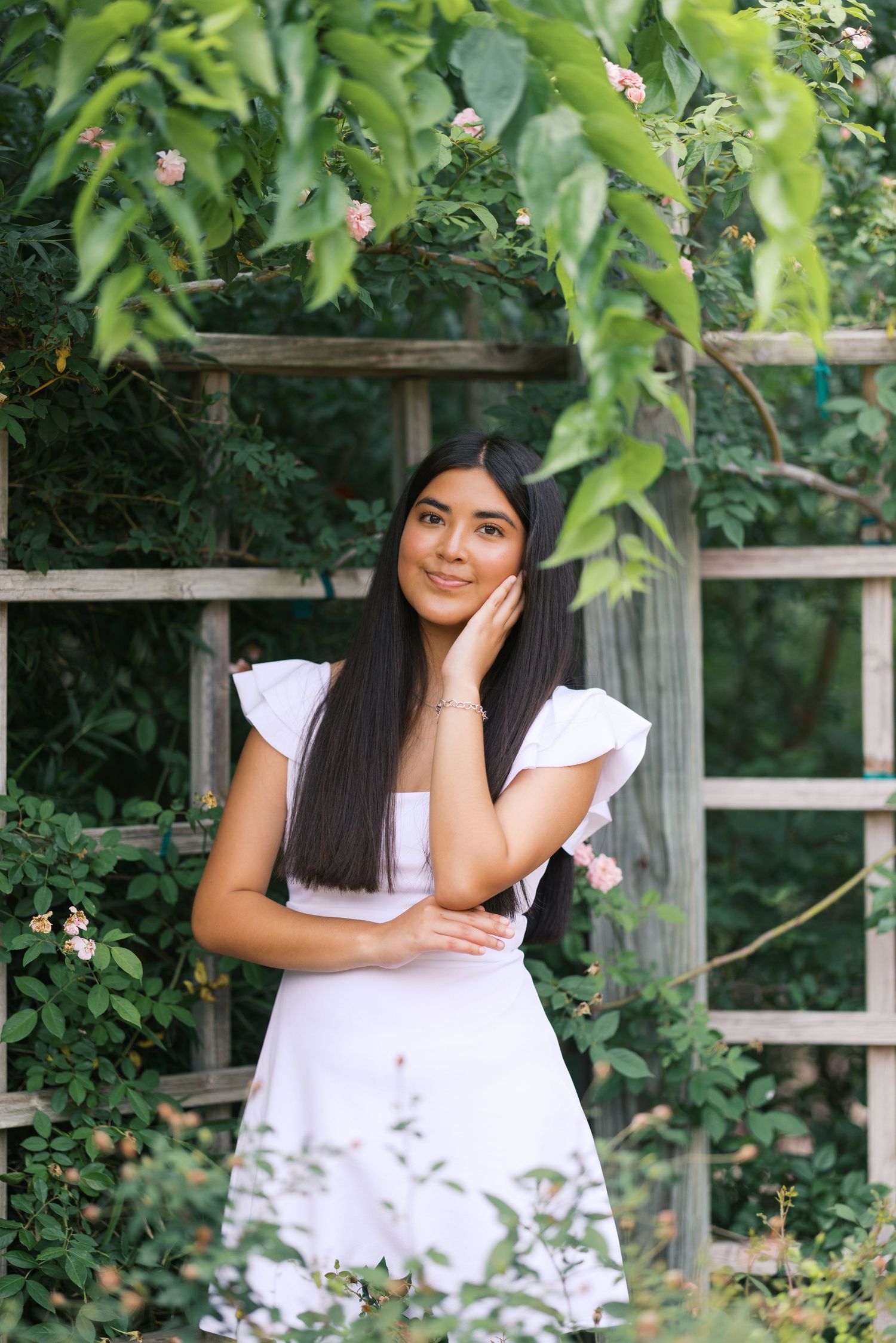 Young woman in white dress poses gracefully in a lush garden with wooden trellis and blooming roses.