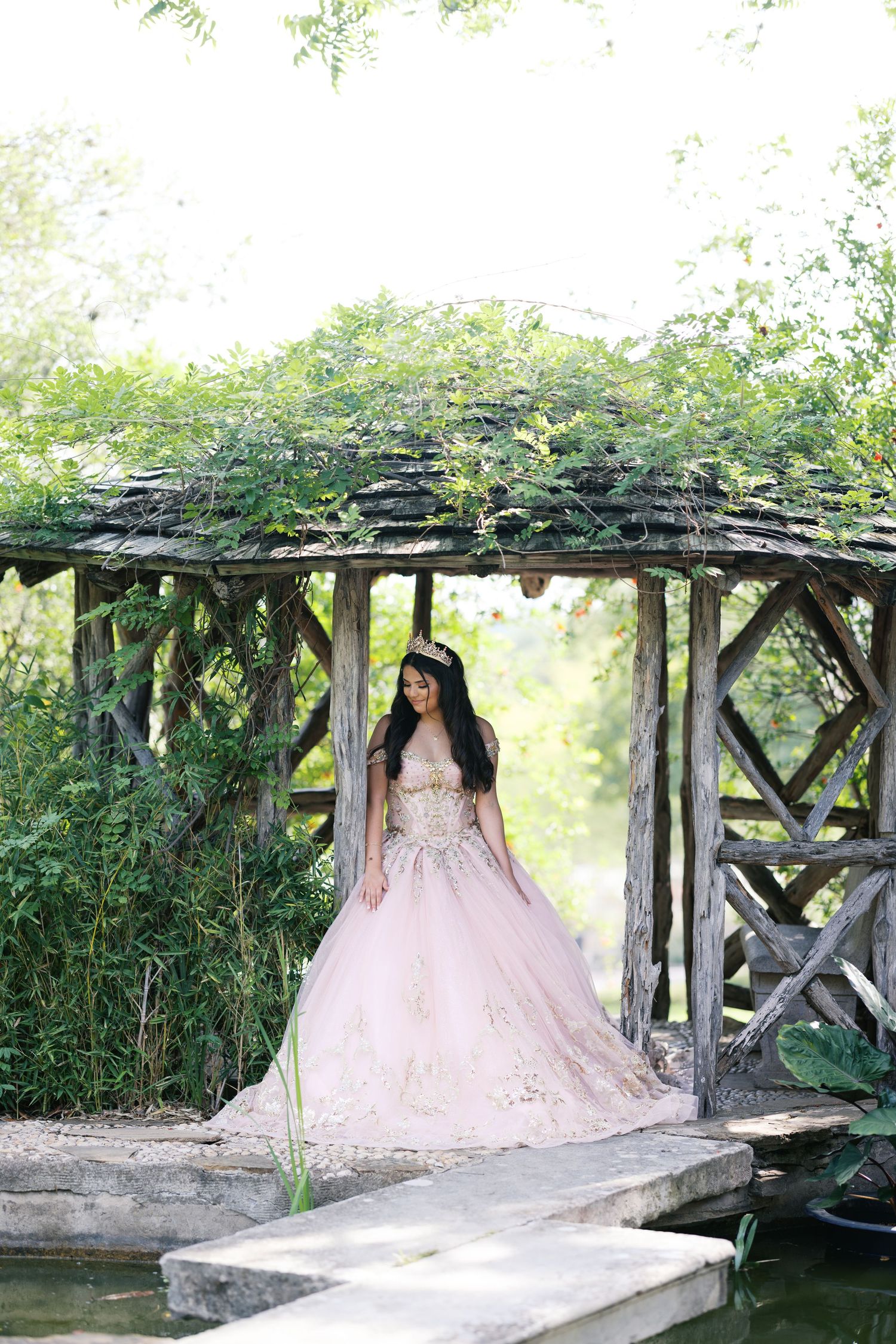 Young woman in pink ball gown and tiara posing under rustic wooden garden arbor covered in lush greenery.