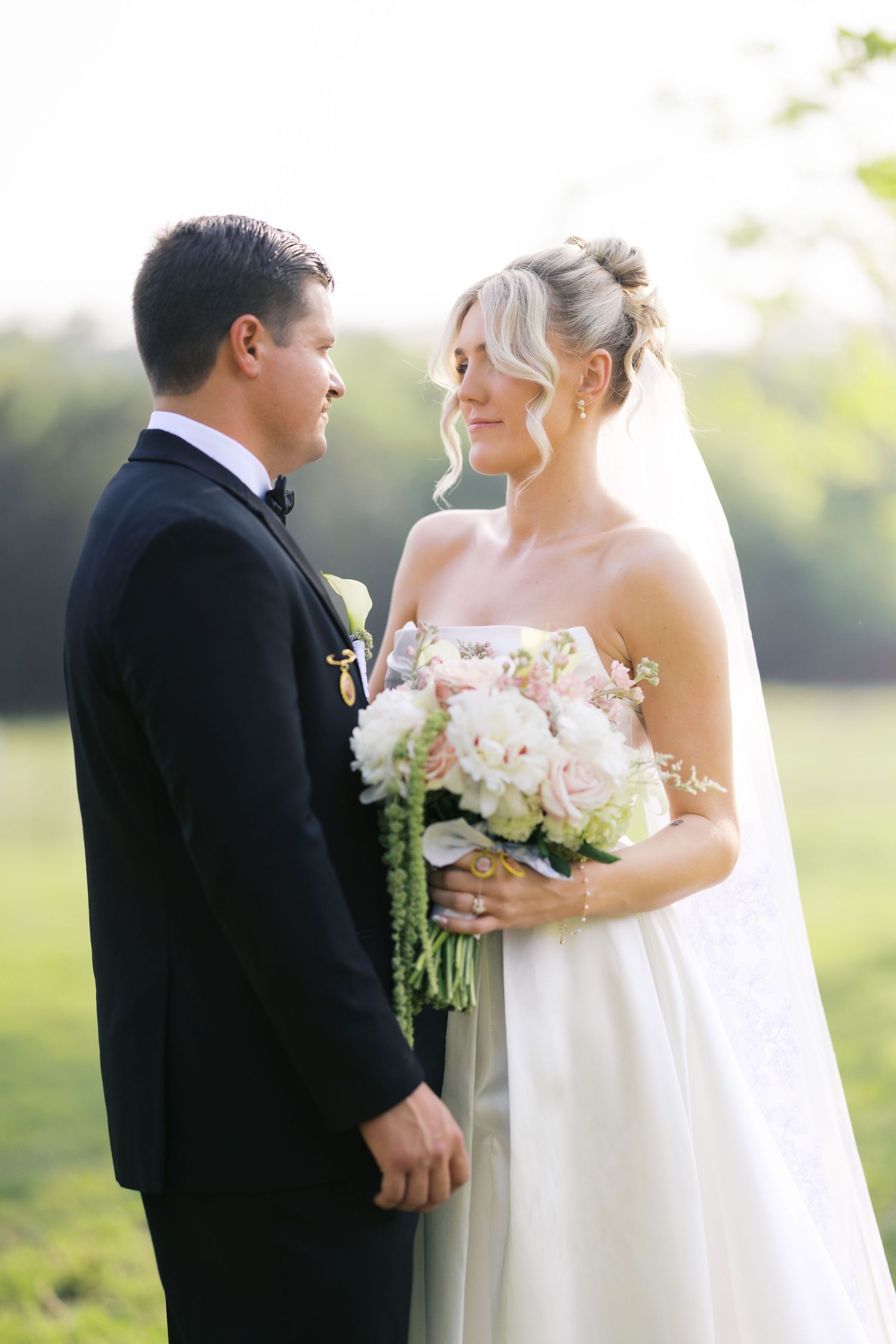 A bride and groom share a tender moment outdoors, bride holding a bouquet of pink roses.