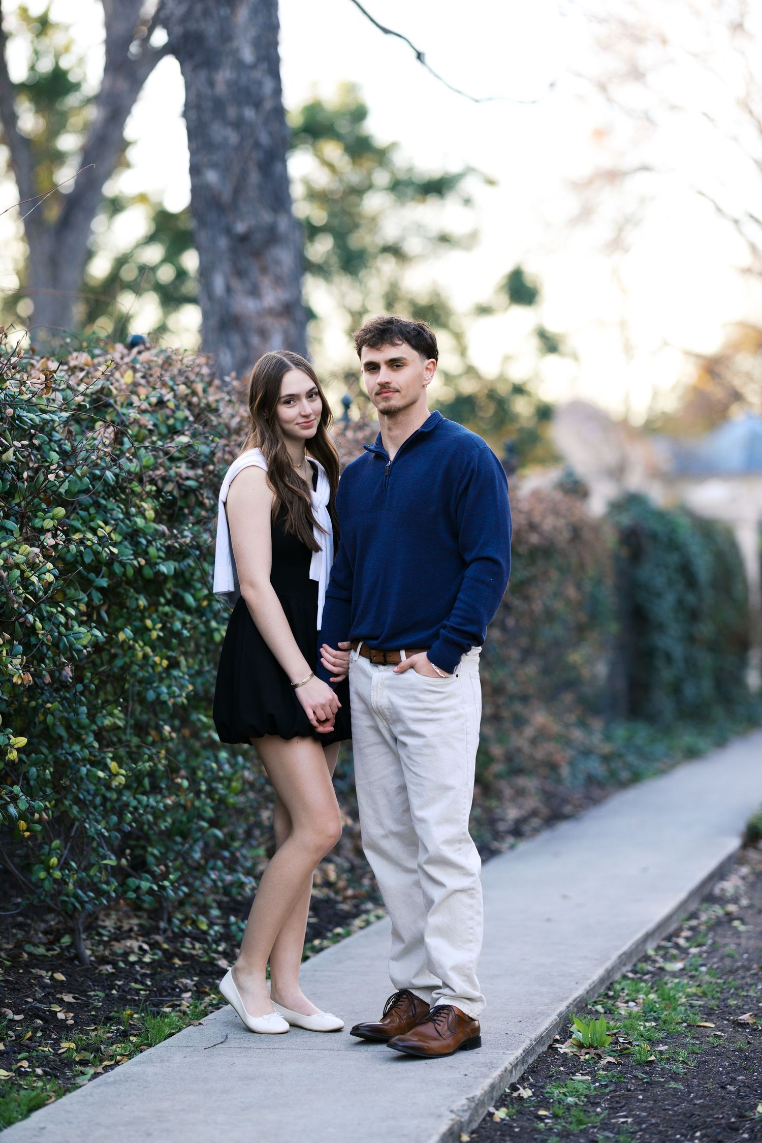 Young couple holding hands on sidewalk beside lush green hedges in outdoor park setting.