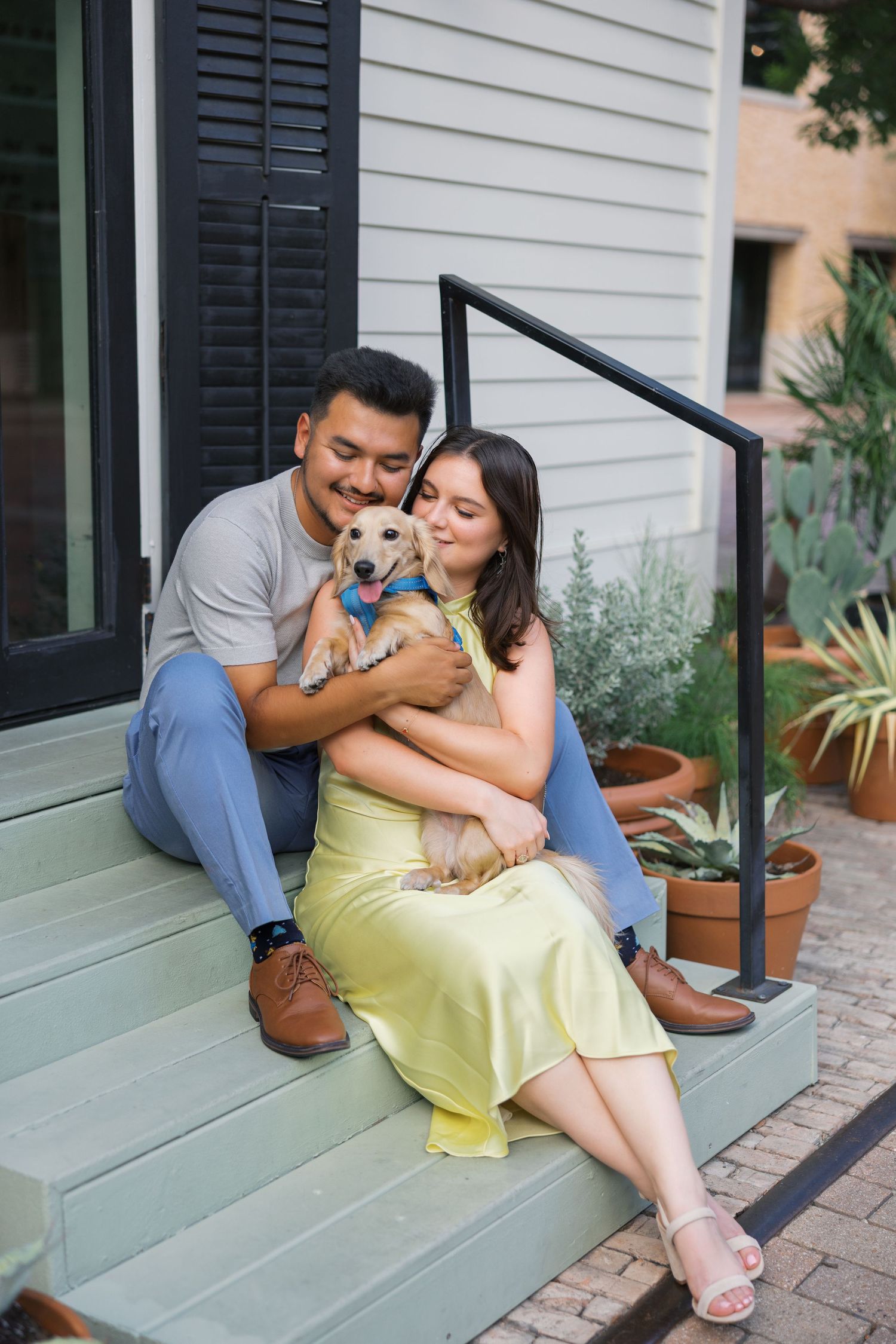 A couple sits on porch steps with their small dog while surrounded by potted plants.