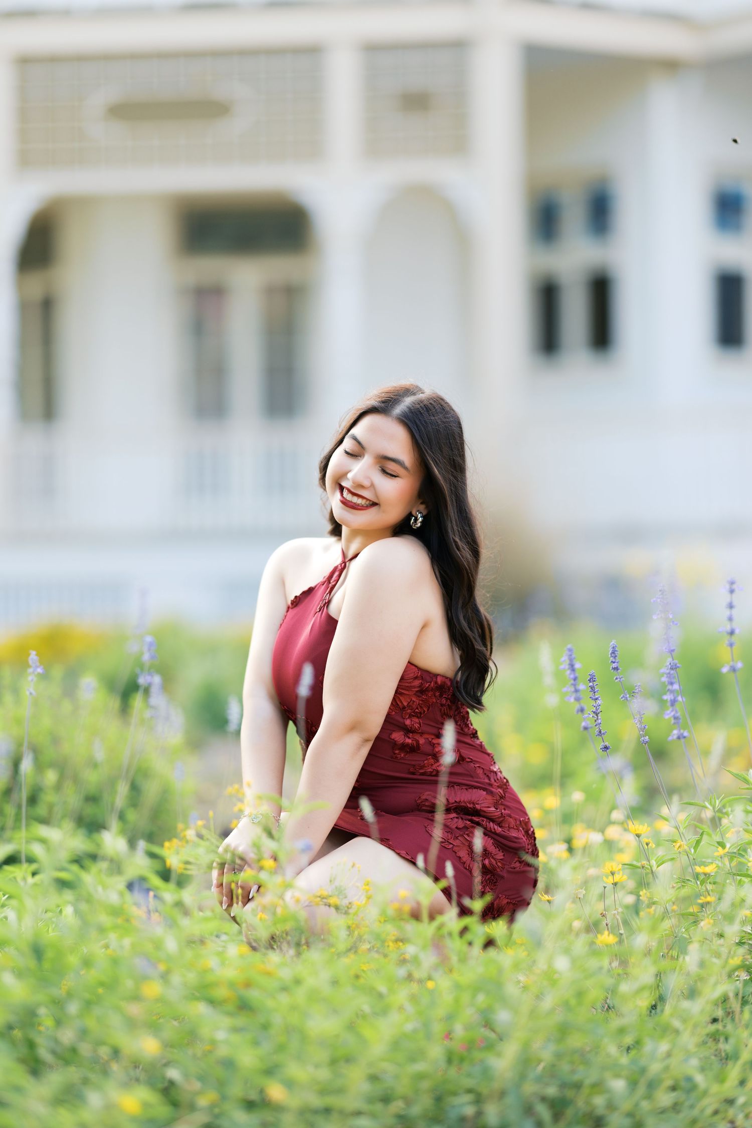 Young woman in burgundy dress sitting among wildflowers, smiling joyfully with a white building in the background.