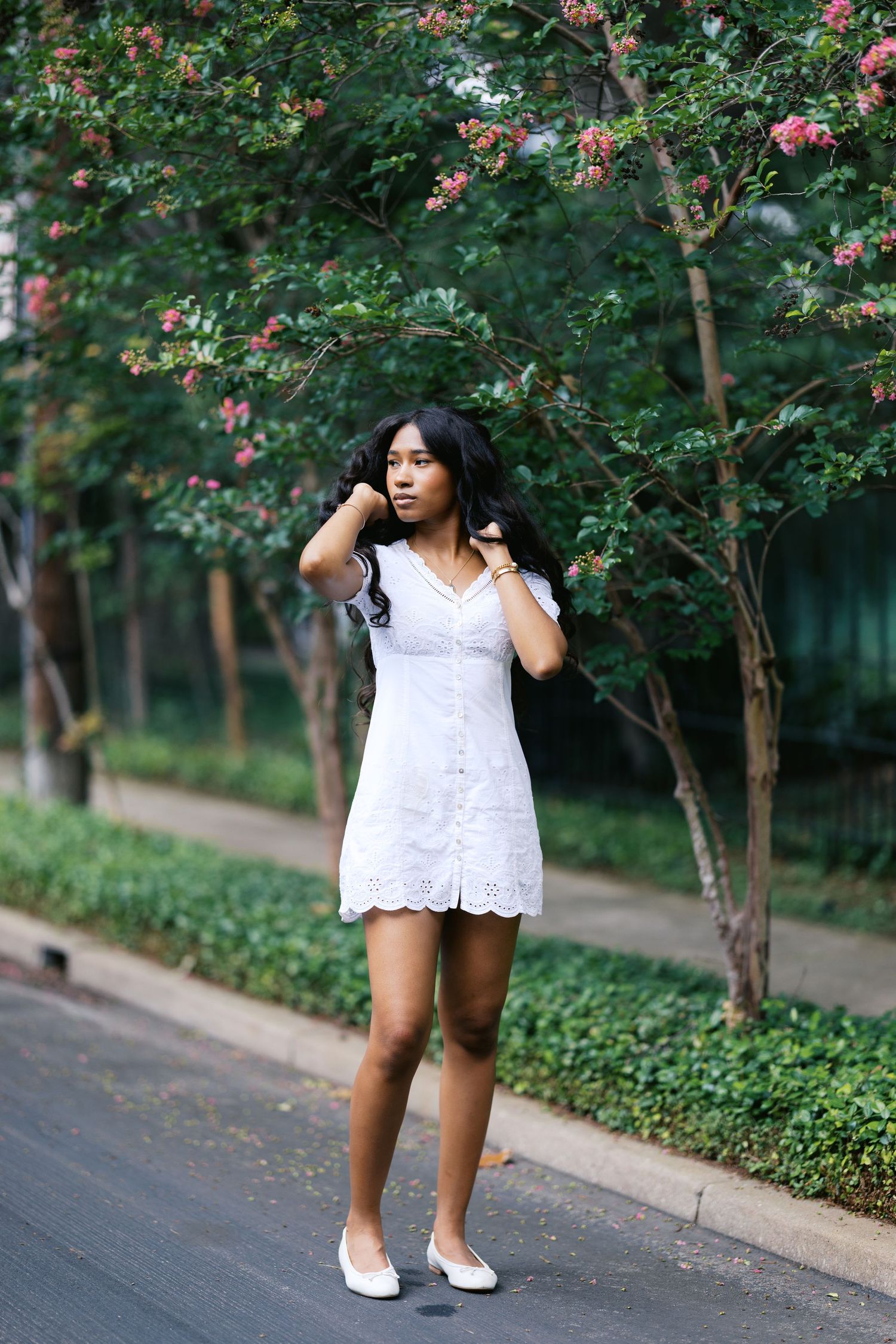 Young woman in white lace mini dress posing outdoors among green trees with pink flowers.
