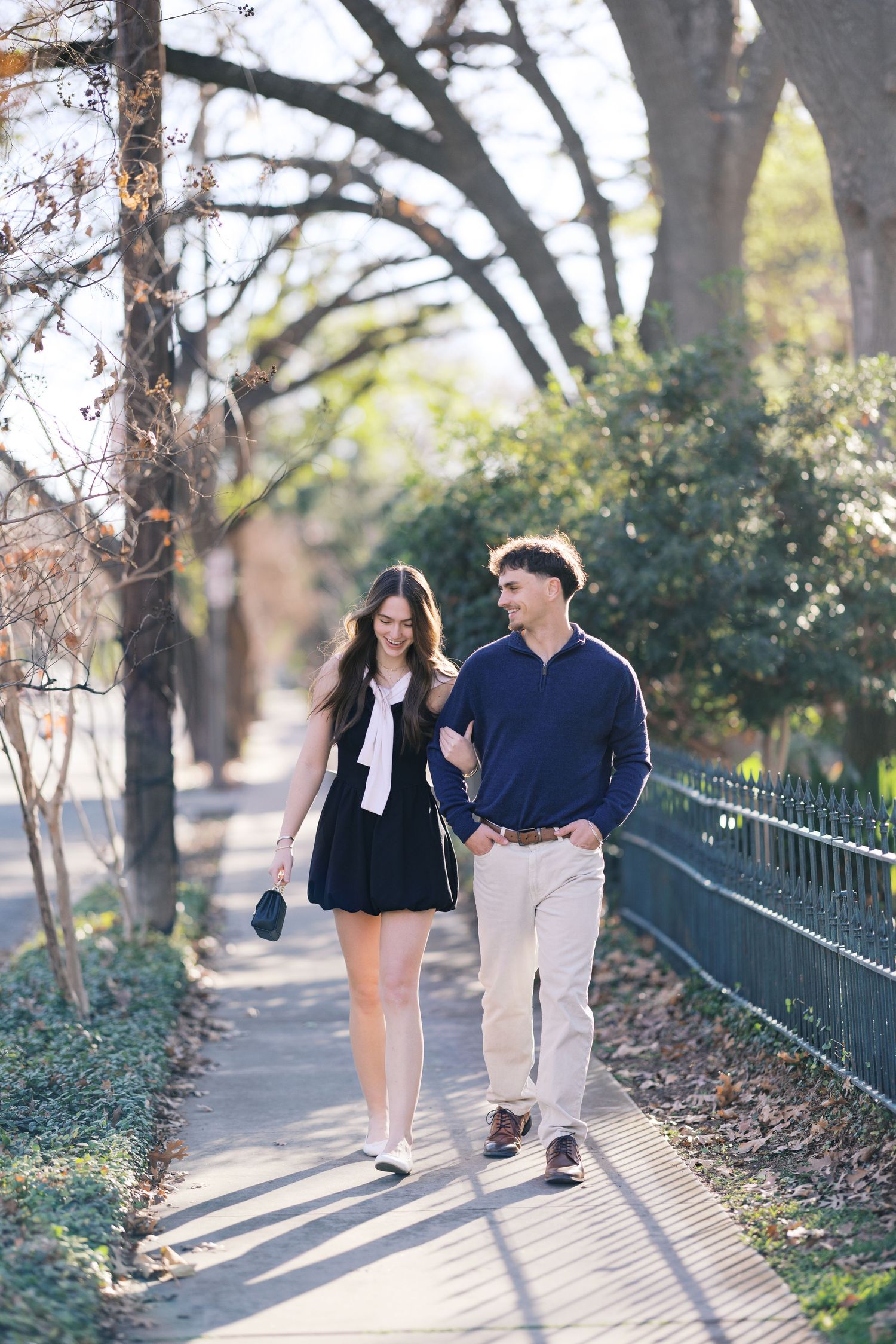 A couple walks hand-in-hand along a sunny tree-lined path, smiling at each other, dressed in navy and khaki.