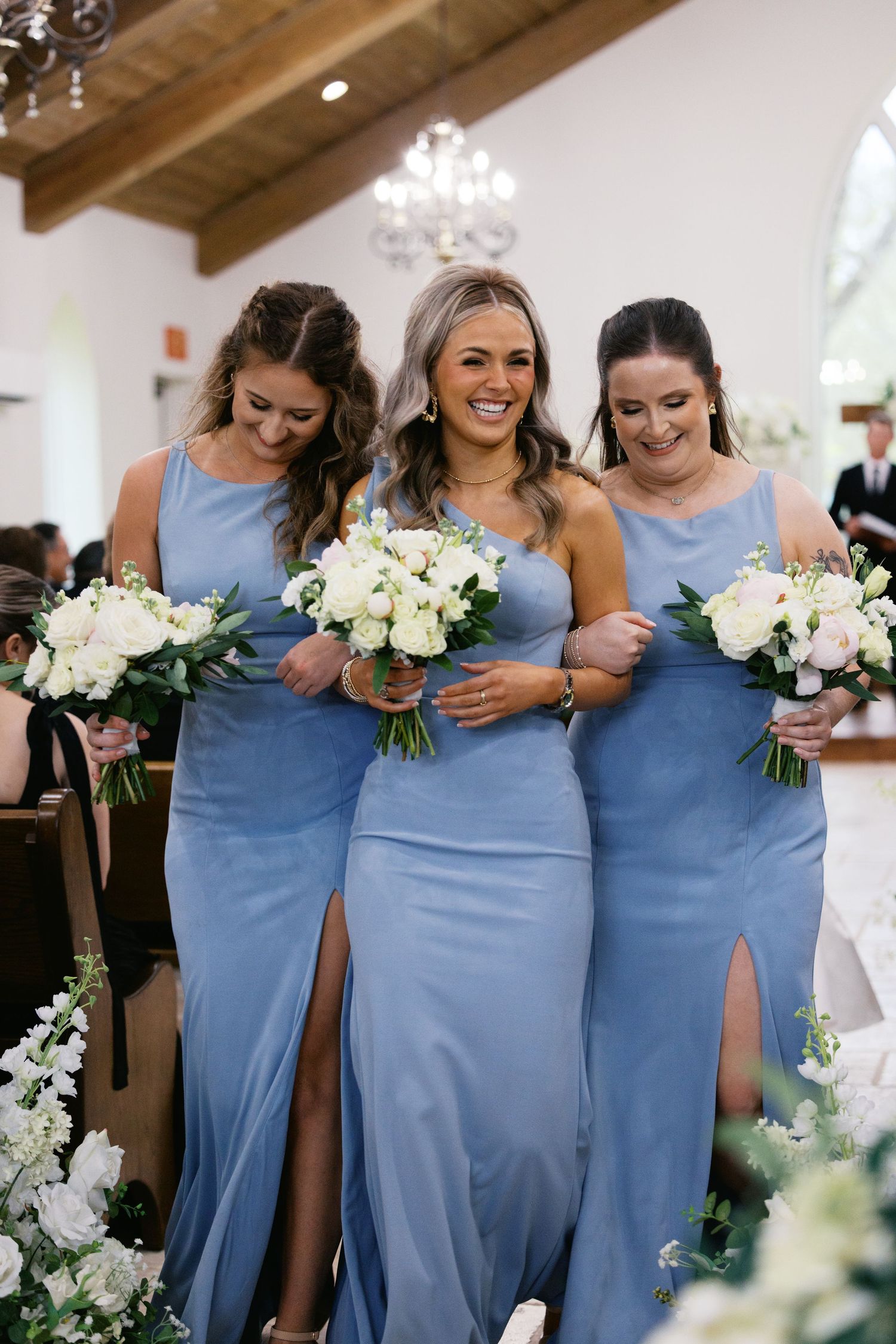 Three bridesmaids in blue satin gowns carrying white bouquets walking down a church aisle smiling.