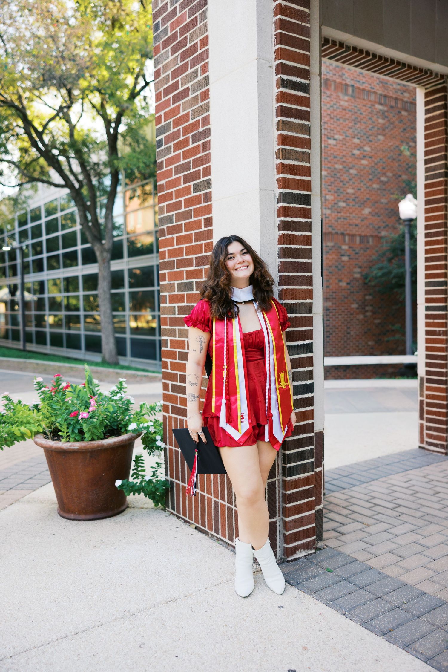 Graduate in academic regalia posing against brick building exterior.