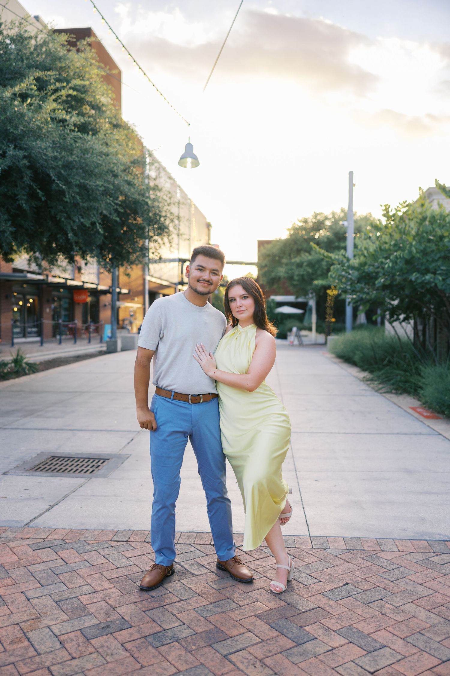 Couple posing on brick walkway with buildings and trees in background.