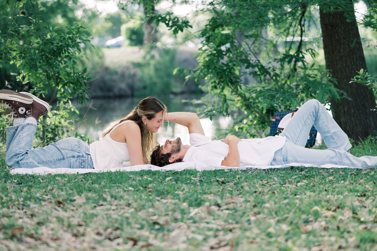A couple enjoys a romantic picnic on a white blanket in a lush green park setting.