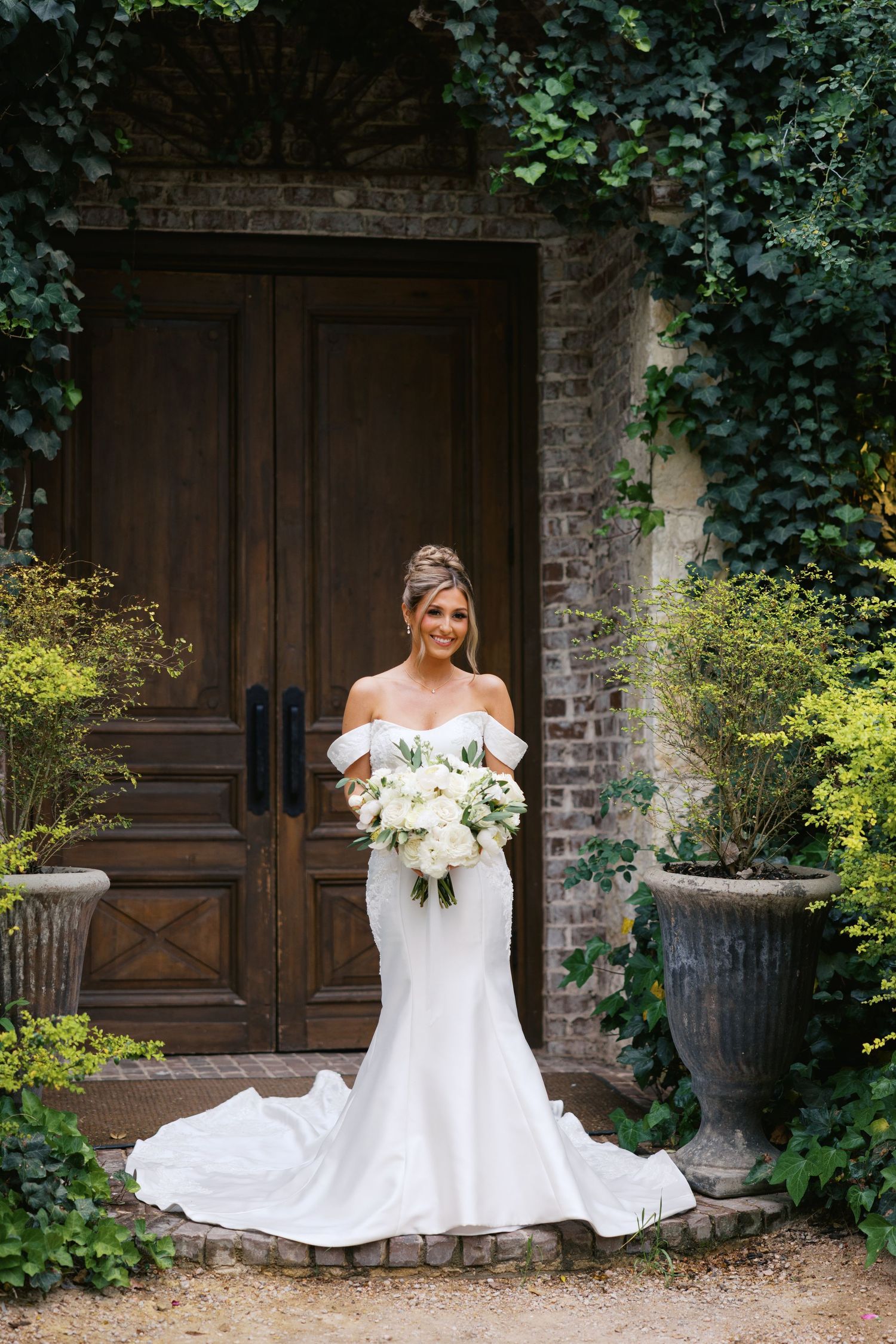 Bride in off-shoulder mermaid gown holds white bouquet before ivy-covered wooden doors.