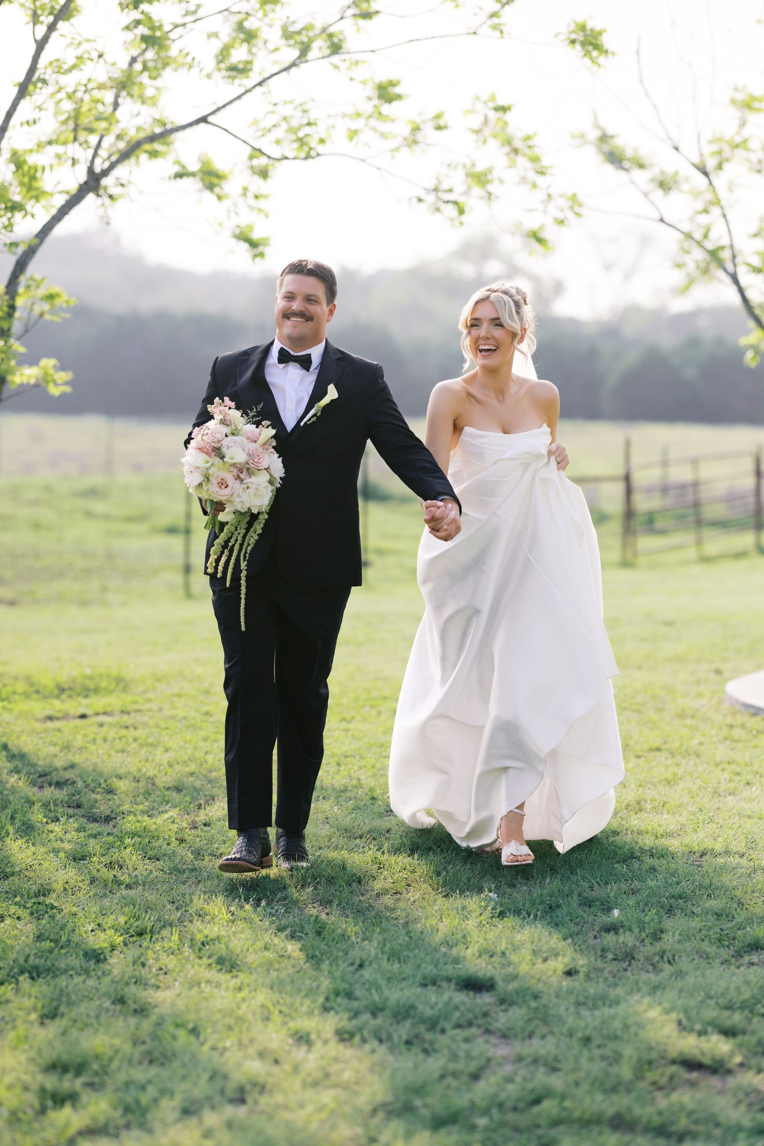 Laughing bride in strapless white gown and groom in black tuxedo walk hand in hand across green meadow.