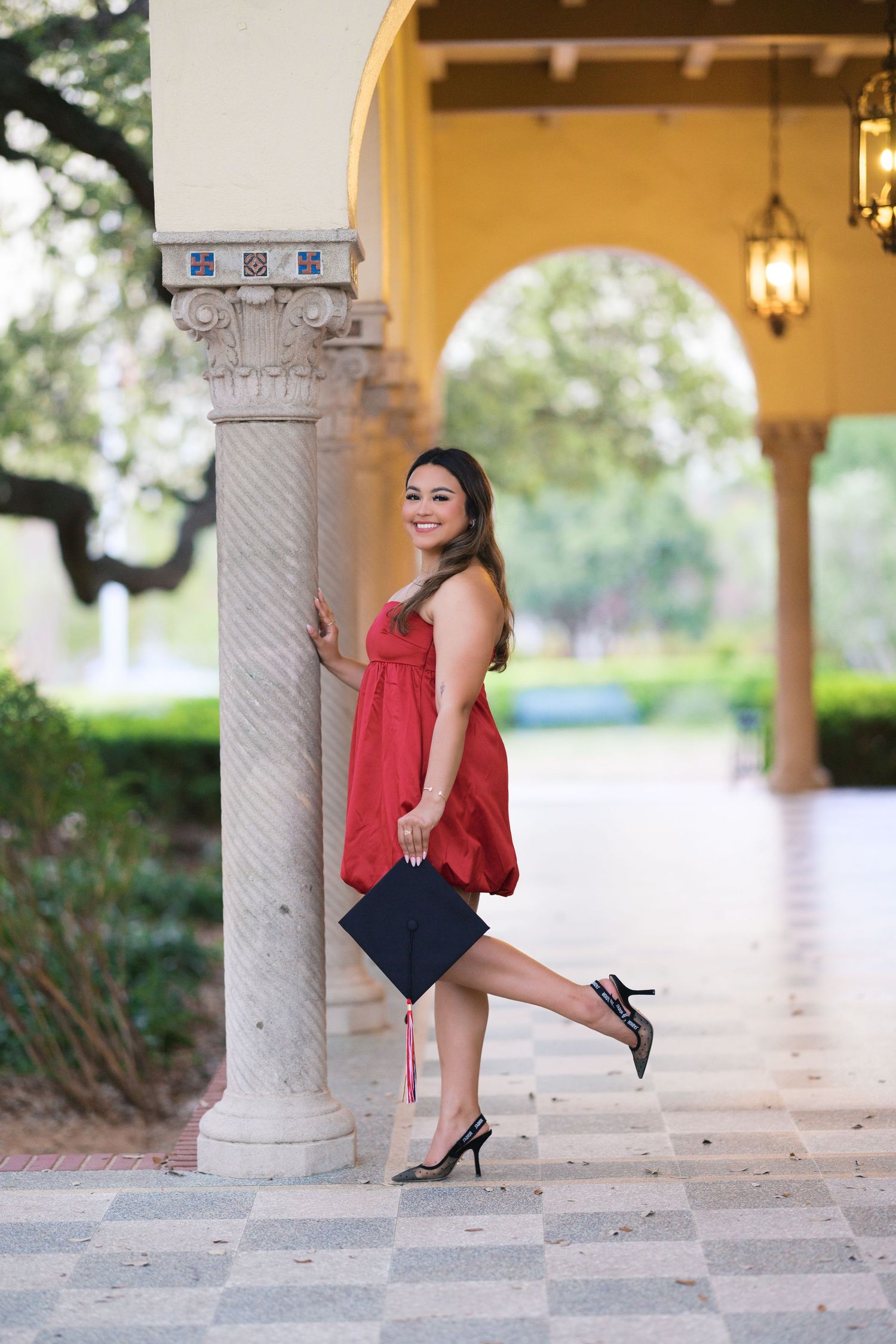Smiling graduate in red dress holding cap leans against marble column under arched corridor with lanterns.