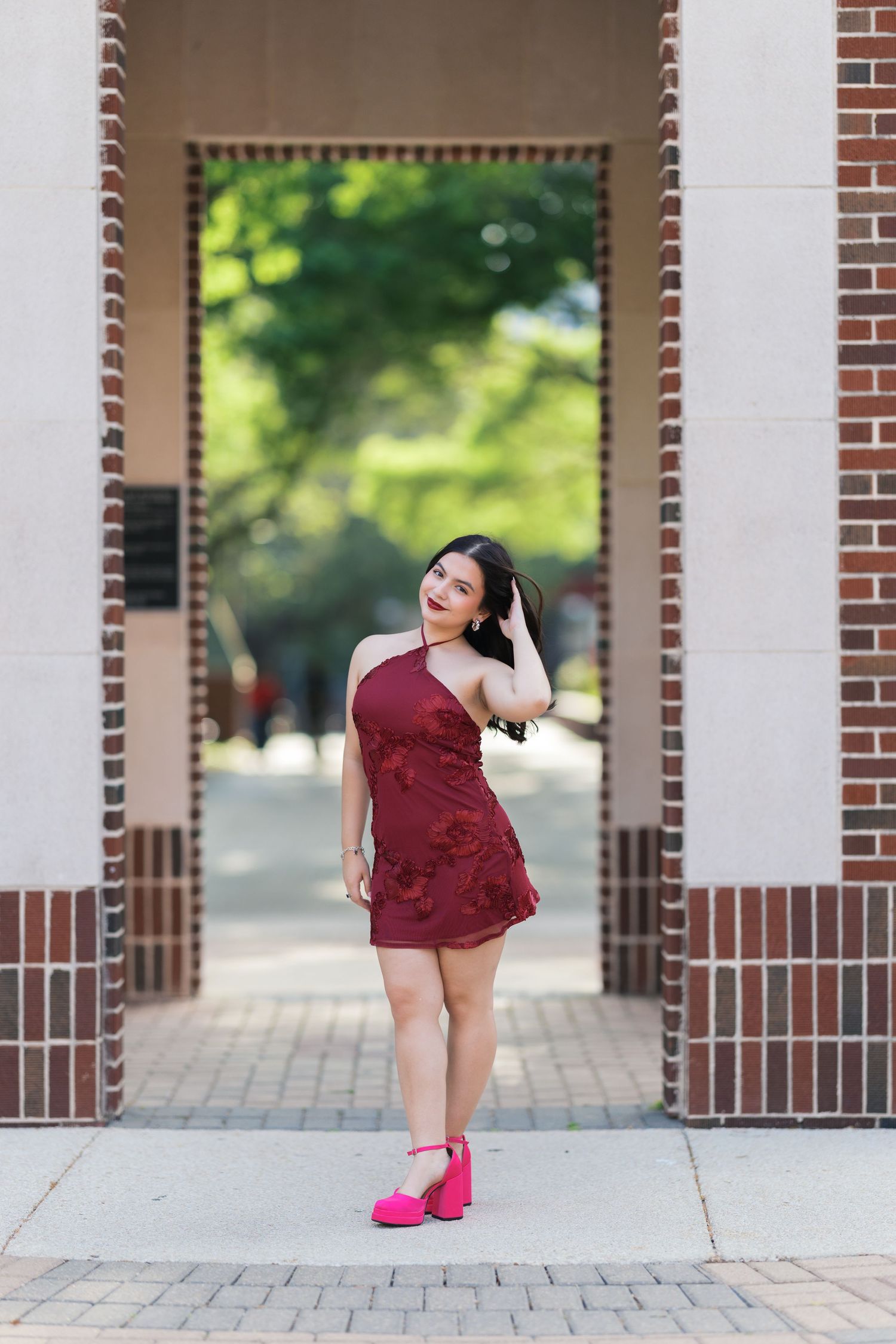 Young woman in burgundy mini dress and pink heels poses under brick archway on sunny day.