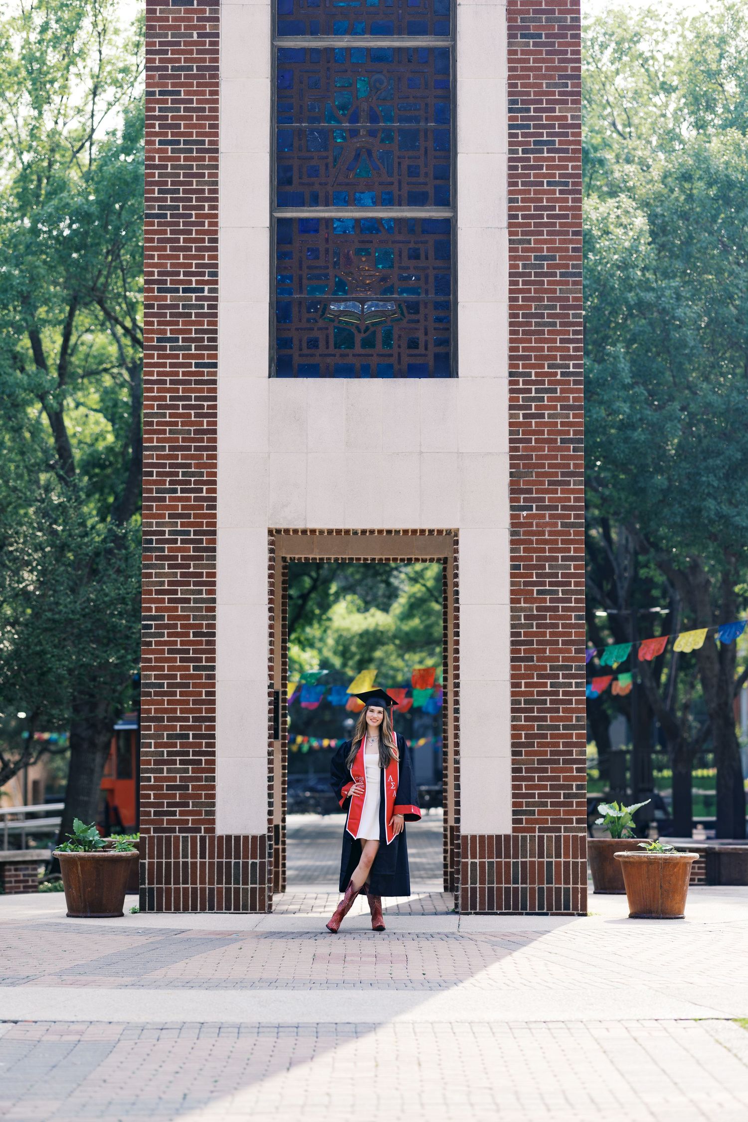 Graduate in cap and gown posing under brick arch with stained glass windows on university campus.