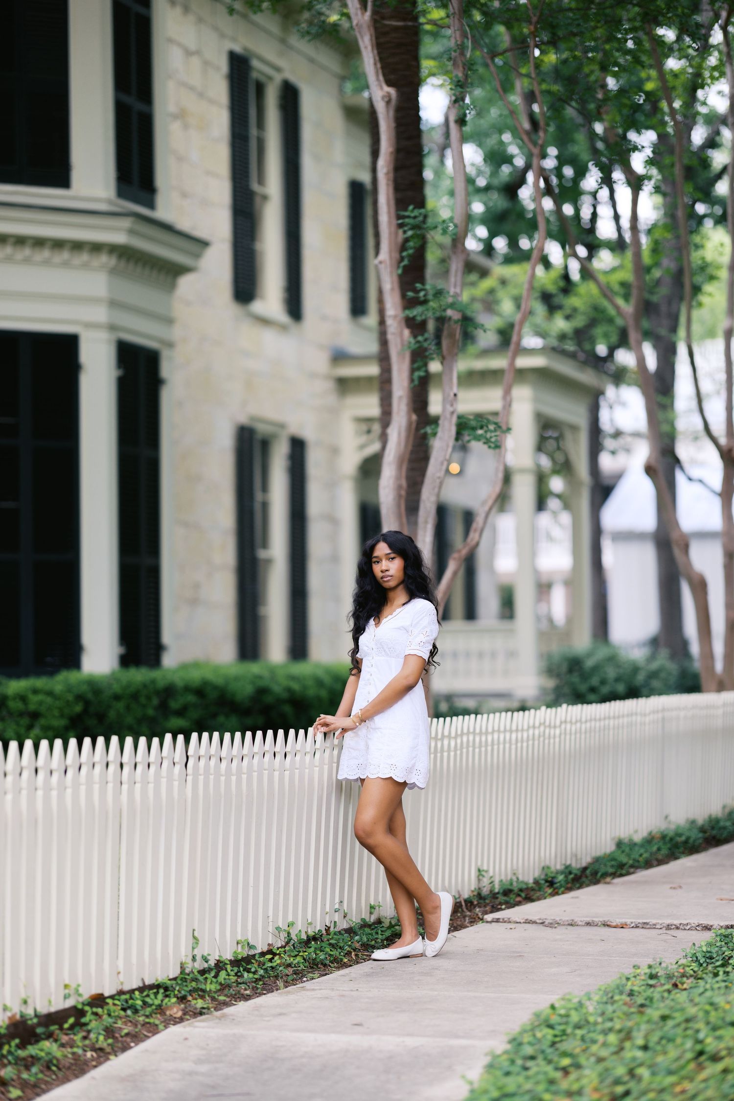 Stylish woman in white eyelet dress leaning against white picket fence near historic home.