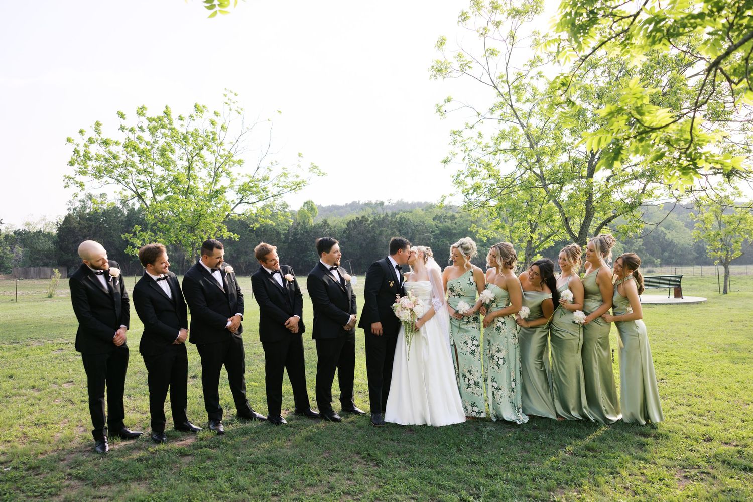 Wedding party posing outdoors with bride and groom kissing, groomsmen in black tuxedos and bridesmaids in sage green gowns.